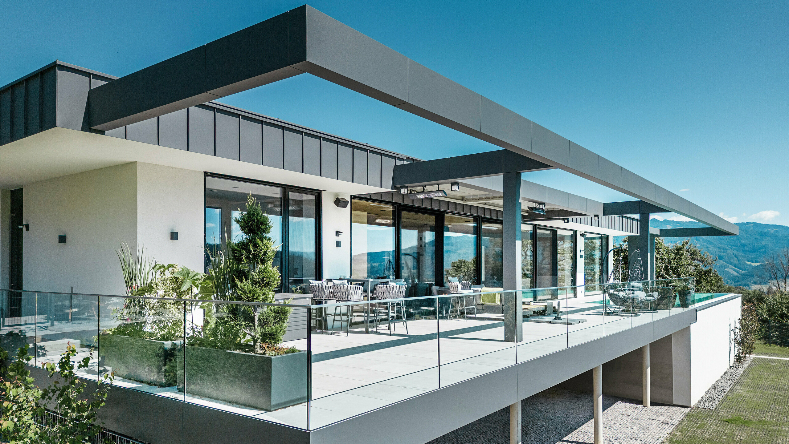 Luxusná terasa rodinného domu v rakúskom Pogieri s elegantnými strešnými a fasádnymi systémami od PREFA. Kombináciou PREFALZ a PREFABOND v tmavošedej farbe vzniká moderný a sofistikovaný dizajn. Priestranná terasa ponúka úchvatný výhľad a je vybavená skleneným zábradlím, ktoré vytvára otvorenú a príjemnú atmosféru. Čiastočne zastrešená vonkajšia plocha vám umožní vychutnať si krásu okolitej prírody a je ideálna na oddych a spoločenské stretnutia.