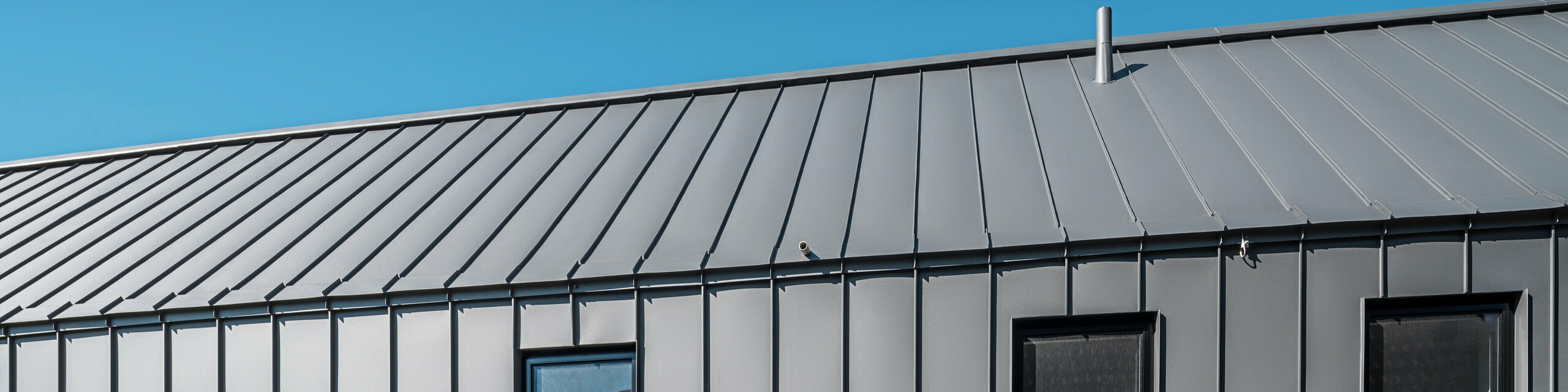 Bočný pohľad na rodinný dom v rakúskom Pogieri s odolným a elegantným zvitkovým plechom PREFALZ v P.10 tmavošedej na streche a fasáde. Hliníkový obklad so stojatou drážkou ponúka čisté vertikálne línie, ktoré elegantne rámujú okná a vytvárajú moderný vzhľad. Minimalistická architektúra stojí pod jasnou modrou oblohou.