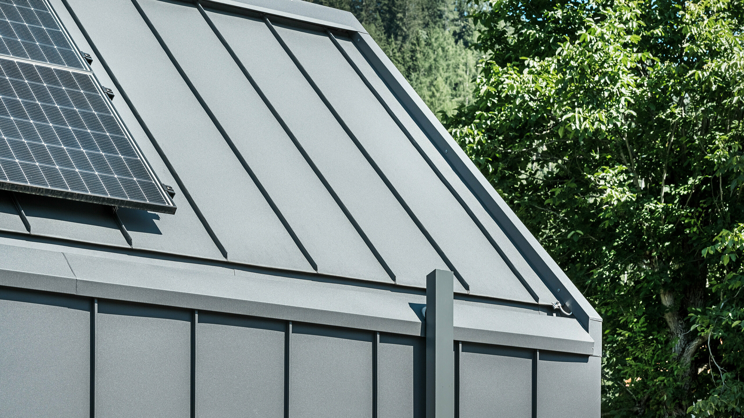 Detailný záber na strechu a fasádu moderného rodinného domu v rakúskom Pogieri s udržateľným a odolným hliníkovým plášťom PREFA. Strecha a fasáda sú pokryté zvitkovým plechom PREFALZ vo farbe P.10 tmavošedá, čo dodáva domu elegantný a nadčasový vzhľad. Dom je navyše vybavený kvalitným odvodnením striech, ktoré dokonale spája funkčnosť a estetiku v architektúre. Odvodnenie strechy prebieha cez hranatú rúru PREFA - tiež v tmavošedej farbe a fotovoltický systém na streche viditeľný na ľavom okraji obrázku podčiarkuje trvalo udržateľný koncept budovy. V pozadí je vidieť zelené stromy a modrú oblohu.