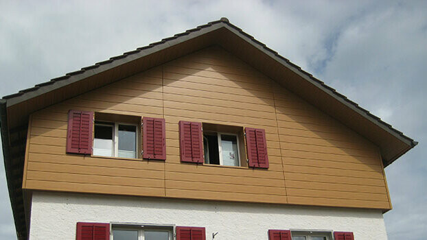 Obloženie štítu klasického domu so sedlovou strechou. Štít je obložený horizontálne montovanými PREFA Sidings v designe svetlého dreva. Okna majú červené okenice.