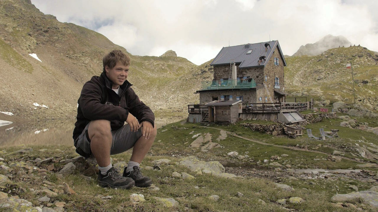 Učeň Jakob Schöttl sedí na kameni. V pozadí vidno chatu „Flagger Schartenhütte“, ktorej strecha bola nanovo zakrytá strešnými panelmi PREFA.