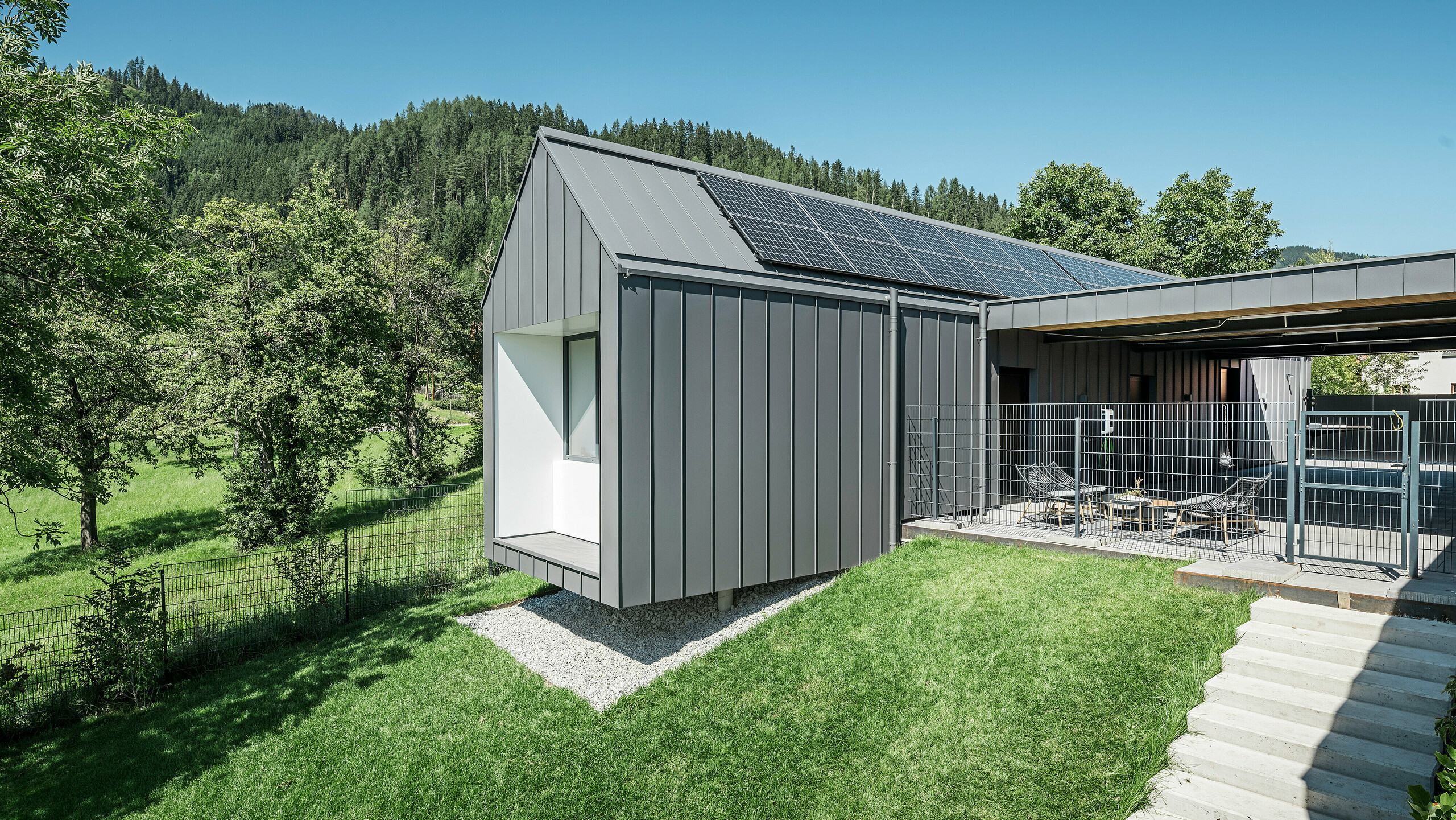 Harmonický zadný pohľad na moderný rodinný dom v rakúskom Pogieri so strešným a fasádnym systémom PREFALZ tmavošedej farby P.10, integrovaným do idylickej lesnej krajiny. Kombinácia tmavošedého hliníkového obkladu a prírodného prostredia podčiarkuje ekologický koncept domu, ktorý dopĺňa efektívny fotovoltický systém na streche. Vonkajší oplotený areál s pohodlným sedením láka k odpočinku a vychutnávaniu si pokoja a sviežosti okolitého lesa.