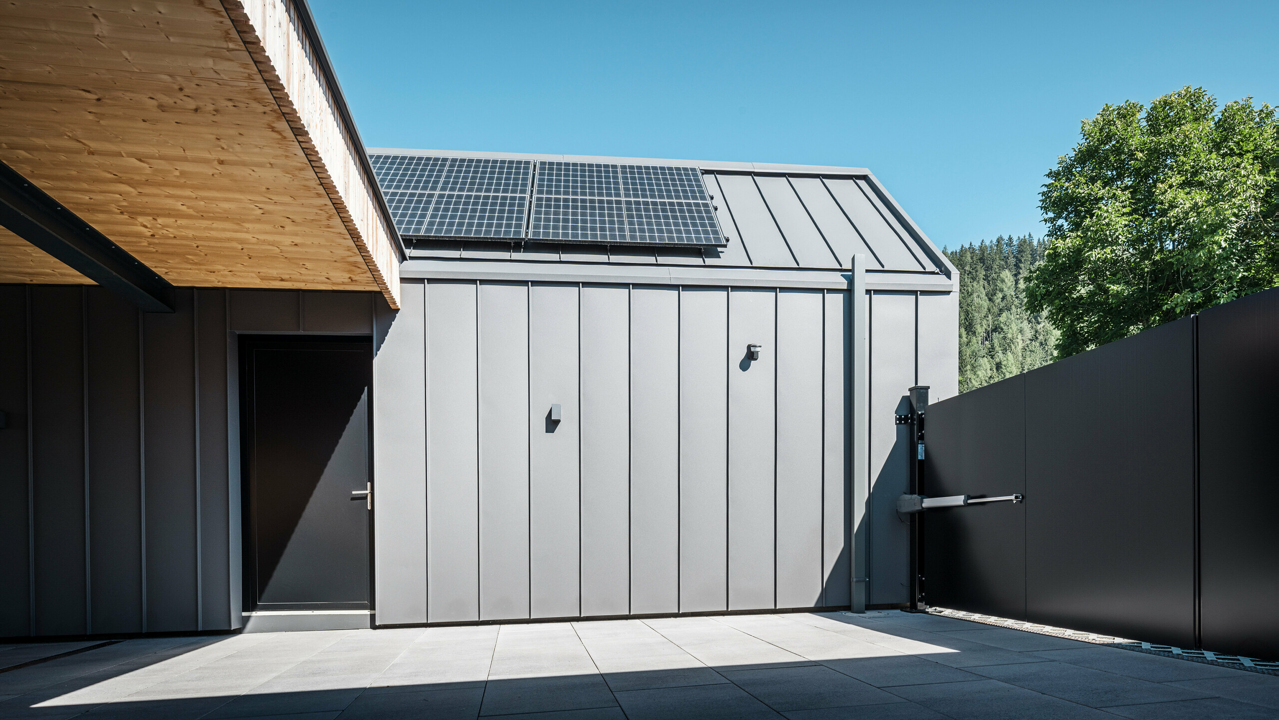 Moderná príjazdová cesta k rodinnému domu v rakúskom Pogieri, ktorý sa vyznačuje jasnými líniami a kvalitnými materiálmi. Plášť budovy je opláštený zvitkovým plechom PREFALZ vo farbe P.10 tmavošedá a tvorí s posuvnou bránou estetický celok. Fotovoltický systém na streche, namontovaný so solárnou nosnou konštrukciou PREFA, podčiarkuje ekologickú orientáciu budovy. Prírodný drevený prelis kontrastuje s moderným kovom a podčiarkuje spojenie tradície a pokroku v architektúre.