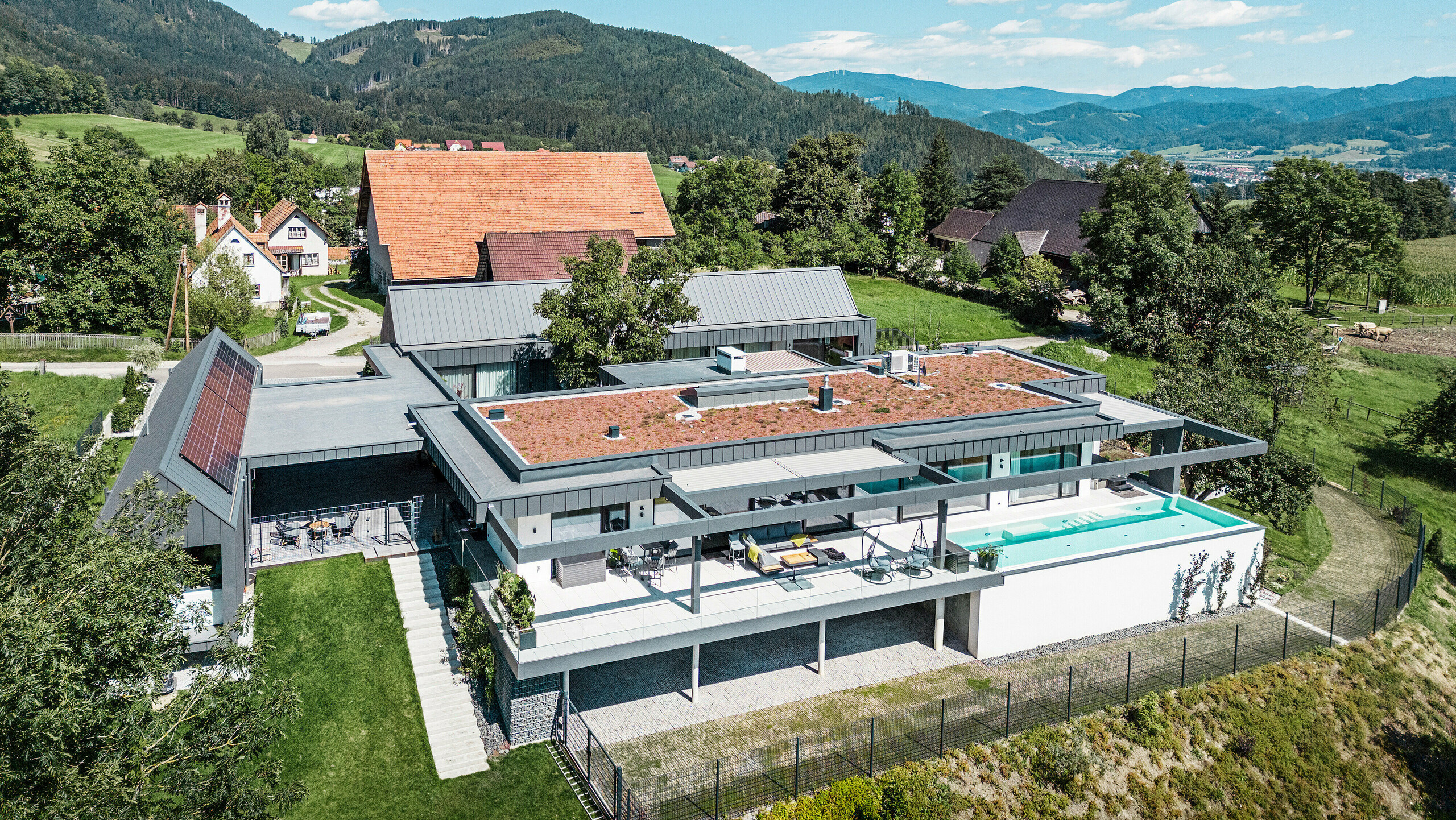 Záber z dronu na pôsobivý rodinný dom s časťami budovy využívanými na poľnohospodárske účely, terasou a bazénom v rakúskom Pogieri. Tmavosivé budovy sú orámované úchvatnou horskou krajinou. Nehnuteľnosť sa prezentuje strešným a fasádnym obkladom PREFALZ v tmavošedej farbe P.10, ktorý podčiarkuje jasné, moderné línie. Terasa a bazén ponúkajú luxusný vonkajší obývací priestor s panoramatickým výhľadom na okolitú krajinu. Spojenie moderných stavebných prvkov z PREFA a idylického miesta vytvára dokonalú symbiózu súčasného dizajnu a prírodných krás.