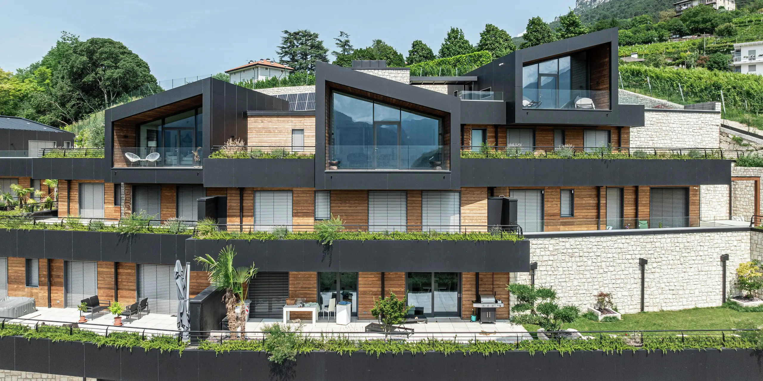 Elegantnosť a funkčnosť v dokonalosti: Výrazná architektúra tohto rezidenčného komplexu sa vyznačuje modernými tvarmi štítov, veľkými sklenenými plochami a harmonickou kombináciou materiálov. Fasáda z hliníkových kompozitných dosiek PREFA v čiernej farbe P.10 zaisťuje odolný a bezúdržbový plášť budovy, zatiaľ čo strecha PREFALZ poskytuje optimálnu ochranu. Drevo a prírodný kameň dodávajú teplo a dokonale zapadajú do zeleného prostredia. Zelené terasy a priestranné obytné priestory vytvárajú jedinečný pocit bývania s najvyšším komfortom a panoramatickým výhľadom.