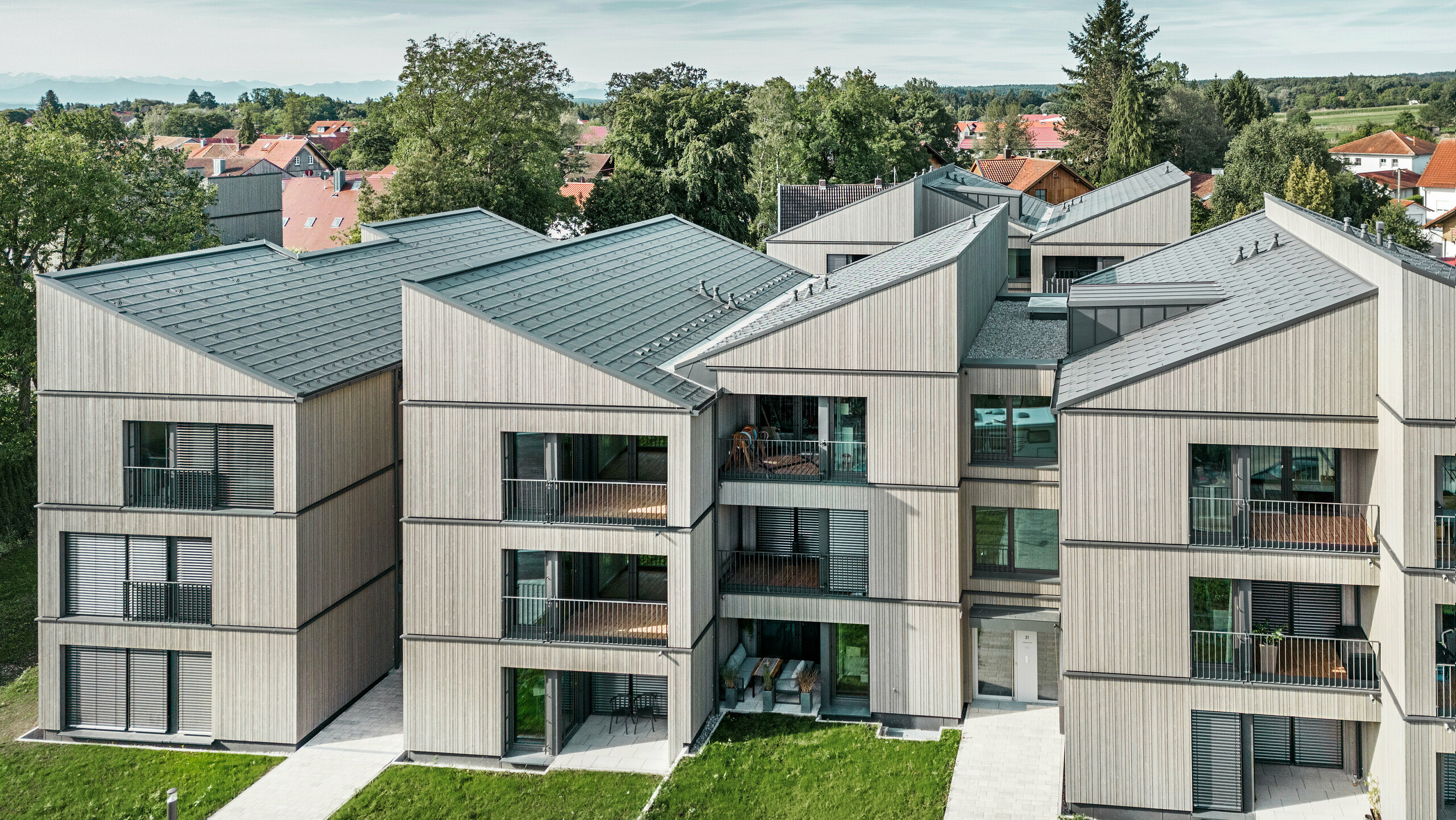 Letecký pohľad na moderný bytový dom na pozemku Schmucker v Utting am Ammersee s udržateľnou hybridnou drevenou konštrukciou a elegantnými drevenými fasádami. Budovy sú vybavené odolnými strešnými panelmi PREFA R.16 v tmavosivej farbe P.10. Túto inovatívnu architektúru, ktorú naplánovala mníchovská architektonická kancelária WWA Architekten, charakterizujú jasné línie a harmonické začlenenie do zeleného okolia. Priestranné balkóny a veľké okná vytvárajú svetlú a priateľskú obytnú atmosféru, zatiaľ čo vysokokvalitné materiály zabezpečujú trvanlivosť a odolnosť voči poveternostným vplyvom.