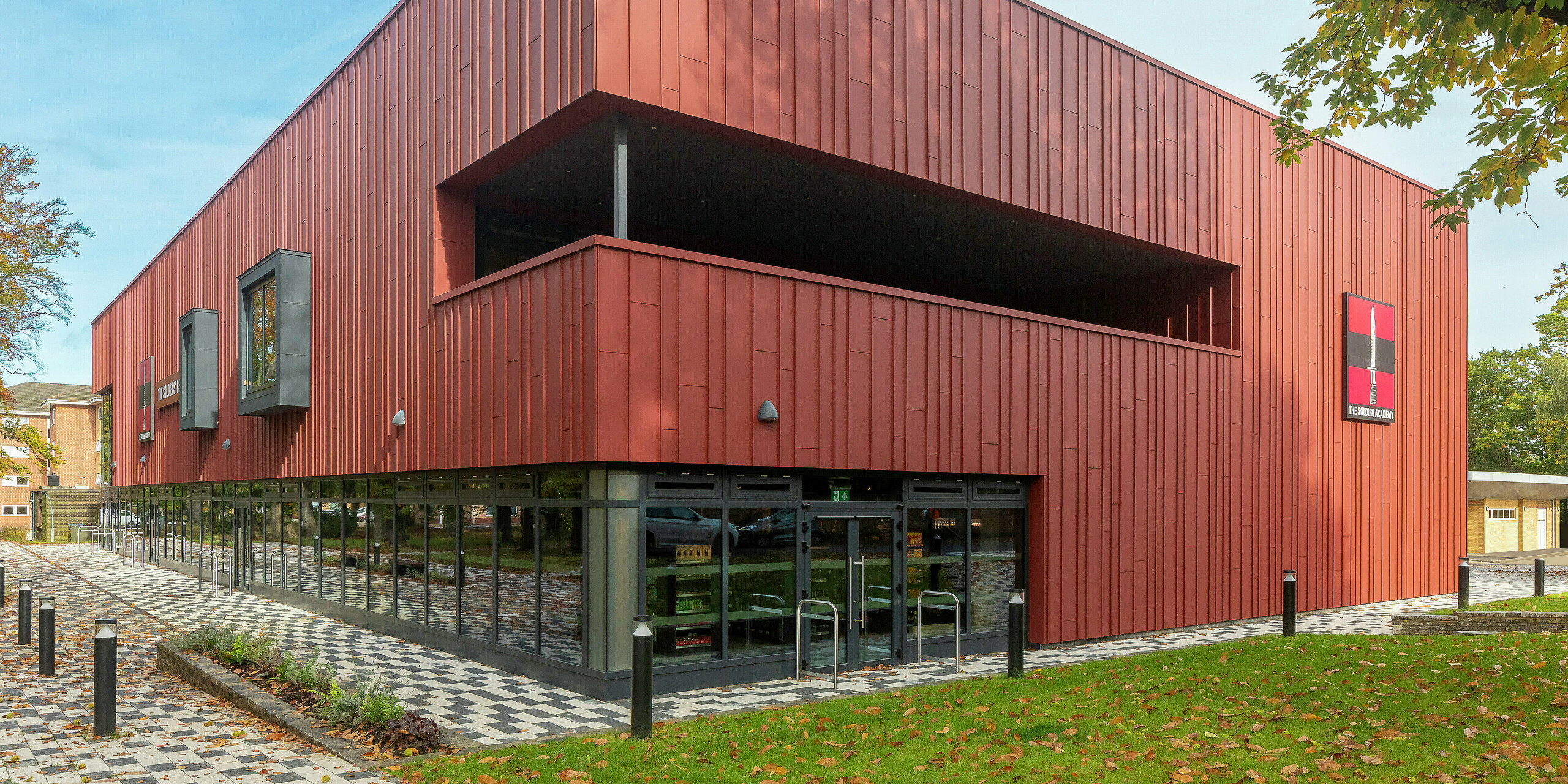 Moderná architektúra s PREFALZ: Soldiers' Centre v Pirbrightu zaujme modernou stojatou falcovou fasádou z PREFALZ v odtieni P.10 tmavočervená. Rozsiahle zasklenie na prízemí vytvára transparentnosť a otvorenosť, zatiaľ čo okenné rámy v P.10 Anthrazit dodávajú štýlové akcenty. Udržateľný stavebný koncept s odolnými hliníkovými produktmi PREFA pre najvyššie nároky na dizajn a kvalitu.