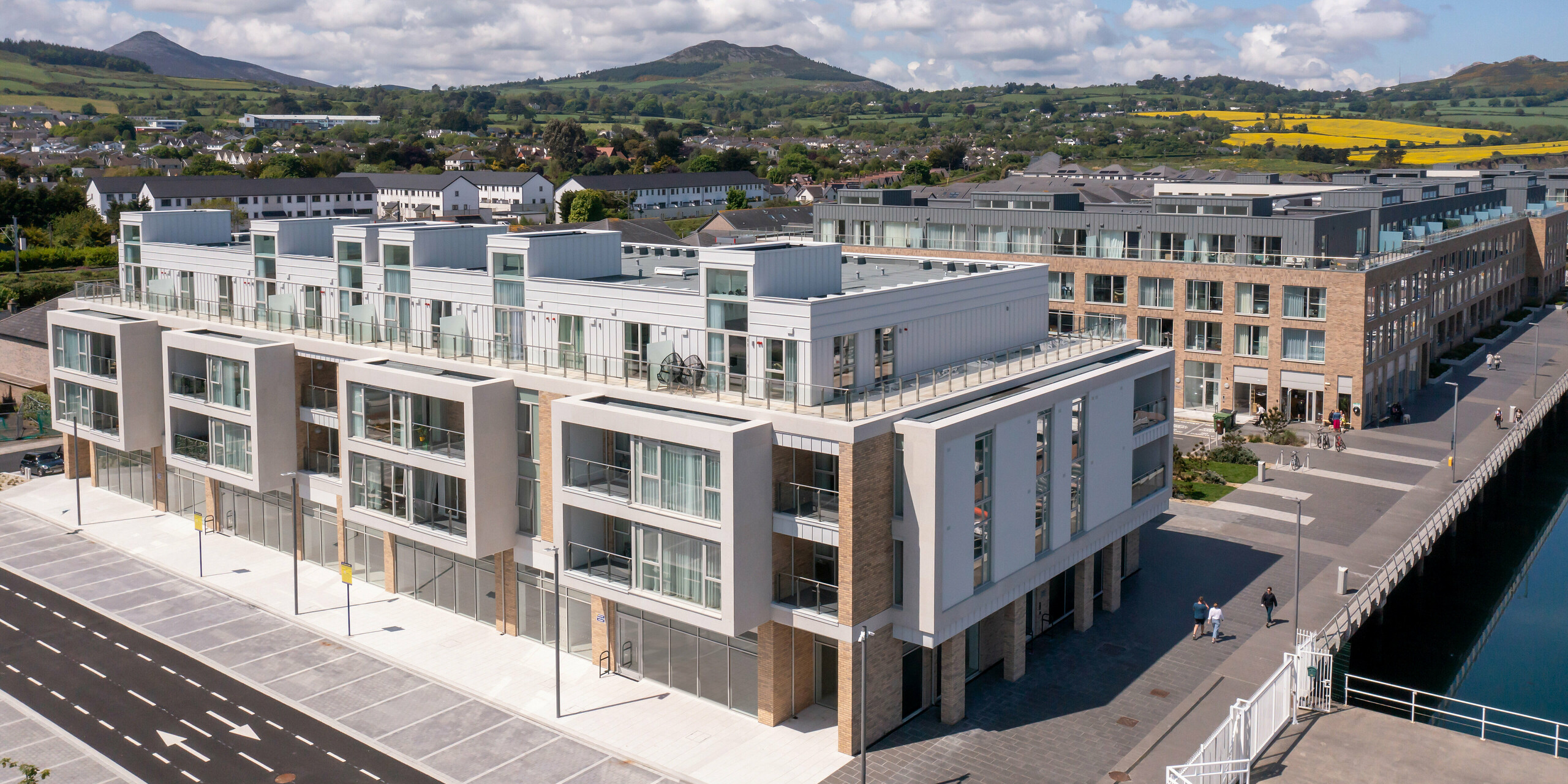 Marina Village Greystones vo Wicklow, Írsko - širokouhlý záber modernej obytnej štvrte priamo na nábreží s výhľadom na prístav. Komplex budov je obložený približne 2 000 m² robustného materiálu PREFALZ vo farbách P.10 prefa biela a P.10 svetlošedá. Elegantná architektúra zahŕňa veľké presklené priečelia, priestranné balkóny a estetický dizajn fasády. Obklopená malebnou krajinou a vysokokvalitným plechom od spoločnosti PREFA ponúka trvalú ochranu a nadčasovú estetiku pre exkluzívne bývanie pri mori.