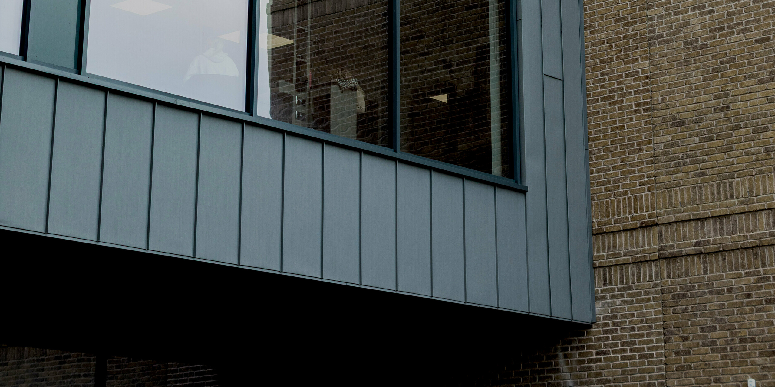 Ein markanter Fassadenausschnitt des Northern Regional College in Ballymena zeigt eine großzügige Glasfront, präzise eingefasst von PREFALZ Aluminium-Stehfalzbahnen in Patinagrau. Die vertikale Struktur des Aluminiums betont die klare Linienführung und rahmt den auskragenden Baukörper elegant ein. Im Kontrast dazu steht das fein strukturierte Klinkermauerwerk im Hintergrund. Die hochwertige PREFA Aluminiumfassade überzeugt durch Langlebigkeit, Witterungsbeständigkeit und eine ruhige, moderne Optik für zeitgemäße Bildungsarchitektur.