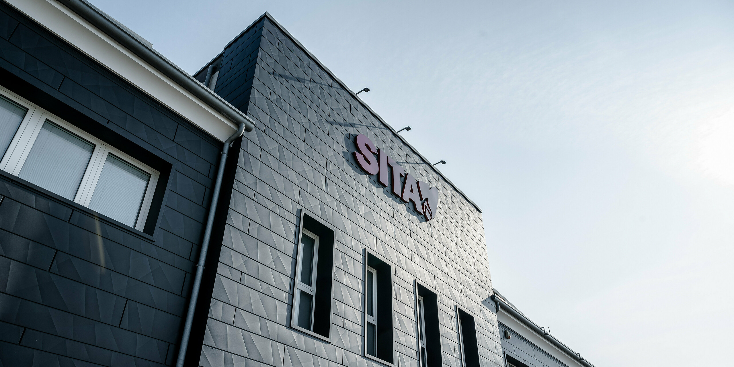 Dynamický pohľad na pôsobivú budovu spoločnosti SITA v talianskom Porcellengu s fasádou PREFA Siding.X v čiernej farbe P.10, ktorá vyniká na modrej oblohe. Fasádne panely odrážajú slnečné svetlo a vytvárajú hru svetla a tieňa, ktorá zdôrazňuje geometrický tvar a kvalitu hliníkového fasádneho systému. Logo spoločnosti vo výraznej červenej farbe tvorí výrazný kontrast ku kovovej fasáde a zdôrazňuje moderný a profesionálny imidž spoločnosti. Tento imidž zdôrazňuje kombináciu funkčnosti a dizajnu, ktorú výrobky PREFA ponúkajú pre súčasné priemyselné budovy. Pod firemným nápisom sa nachádzajú štyri podlhovasté vertikálne okná. Niekoľko okien s bielymi okennými rámami možno rozoznať aj po stranách.