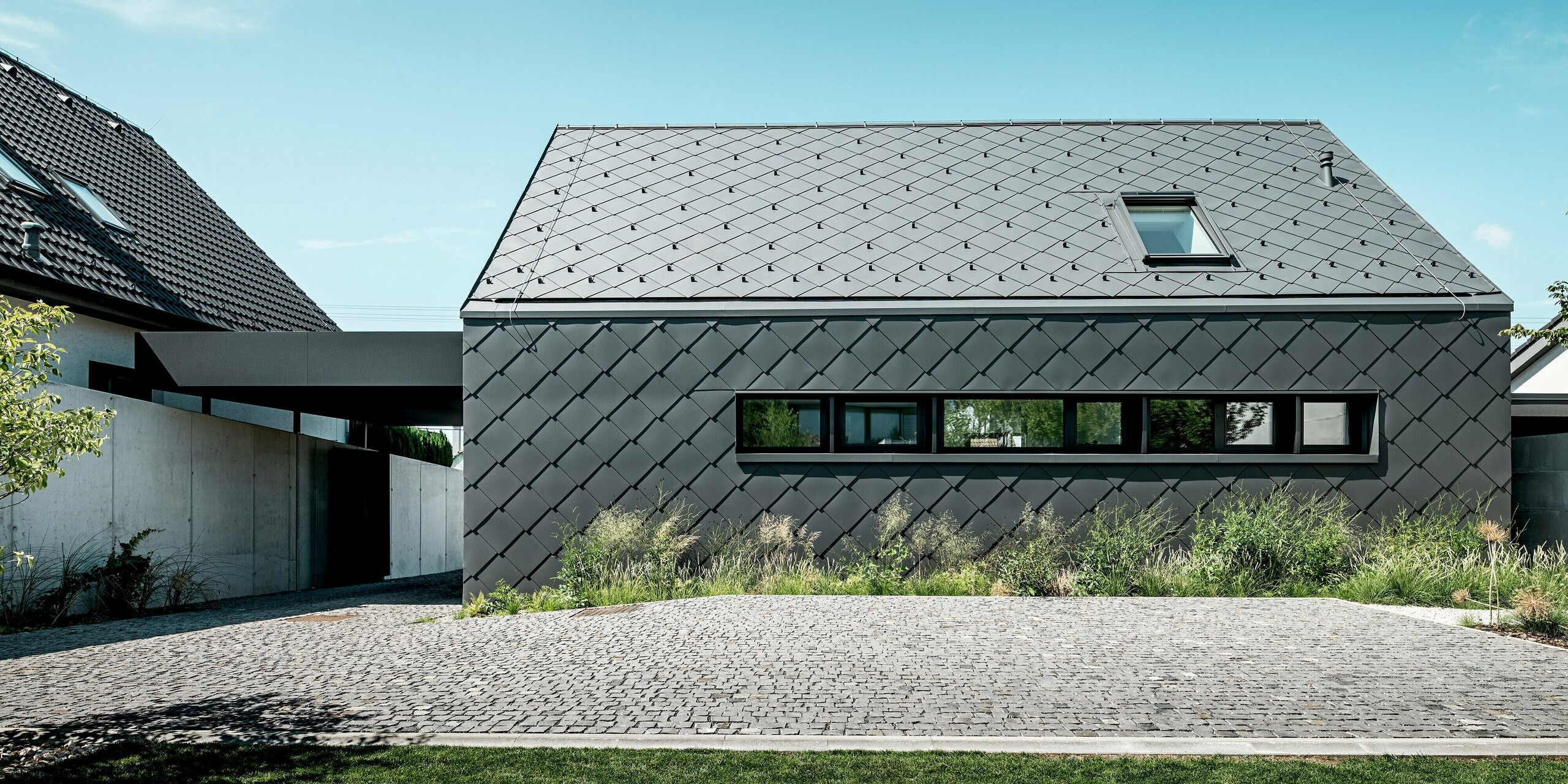 Na obrázku je vidieť jasne štruktúrovaný bočný pohľad na moderný rodinný dom v Trnave, ktorý je kompletne obložený strešnými a fasádnymi šablónami PREFA 44 × 44 v odtieni P.10 tmavošedá. Jednotná štruktúra dodáva streche a fasáde pokojnú, elegantnú rovinnosť a zdôrazňuje precízny tvarový jazyk budovy. Tieto odolné hliníkové produkty ponúkajú vysokú ochranu proti poveternostným vplyvom, farebne stále povrchy a robustný, moderný vzhľad. Prírodná zeleň pred domom vytvára príjemný, jemný kontrast k výraznému kovovému plášťu.