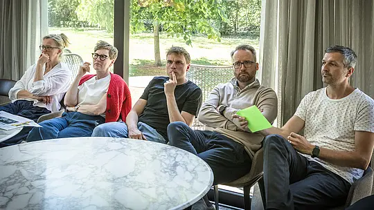 PREFARENZEN 2027 – zasadnutie poroty v svetlej seminárnej miestnosti: Niekoľko architektov a odborníkov na dizajn sedí sústredene v rade pred modrými nástenkami, na ktorých sú prezentované architektonické projekty s hliníkom PREFA. Veľké okná vpúšťajú dovnútra denné svetlo a poskytujú výhľad na zelenú záhradu. Na okrúhlych stoloch ležia knihy o architektúre, zošity a perá. Scéna vyžaruje intenzívnu diskusiu, kreatívne hodnotenie dizajnu a funkčnosti, ako aj inšpiráciu, ktorú hliníkové produkty PREFA ponúkajú prostredníctvom trvácnych, estetických striech a fasád v architektonických projektoch