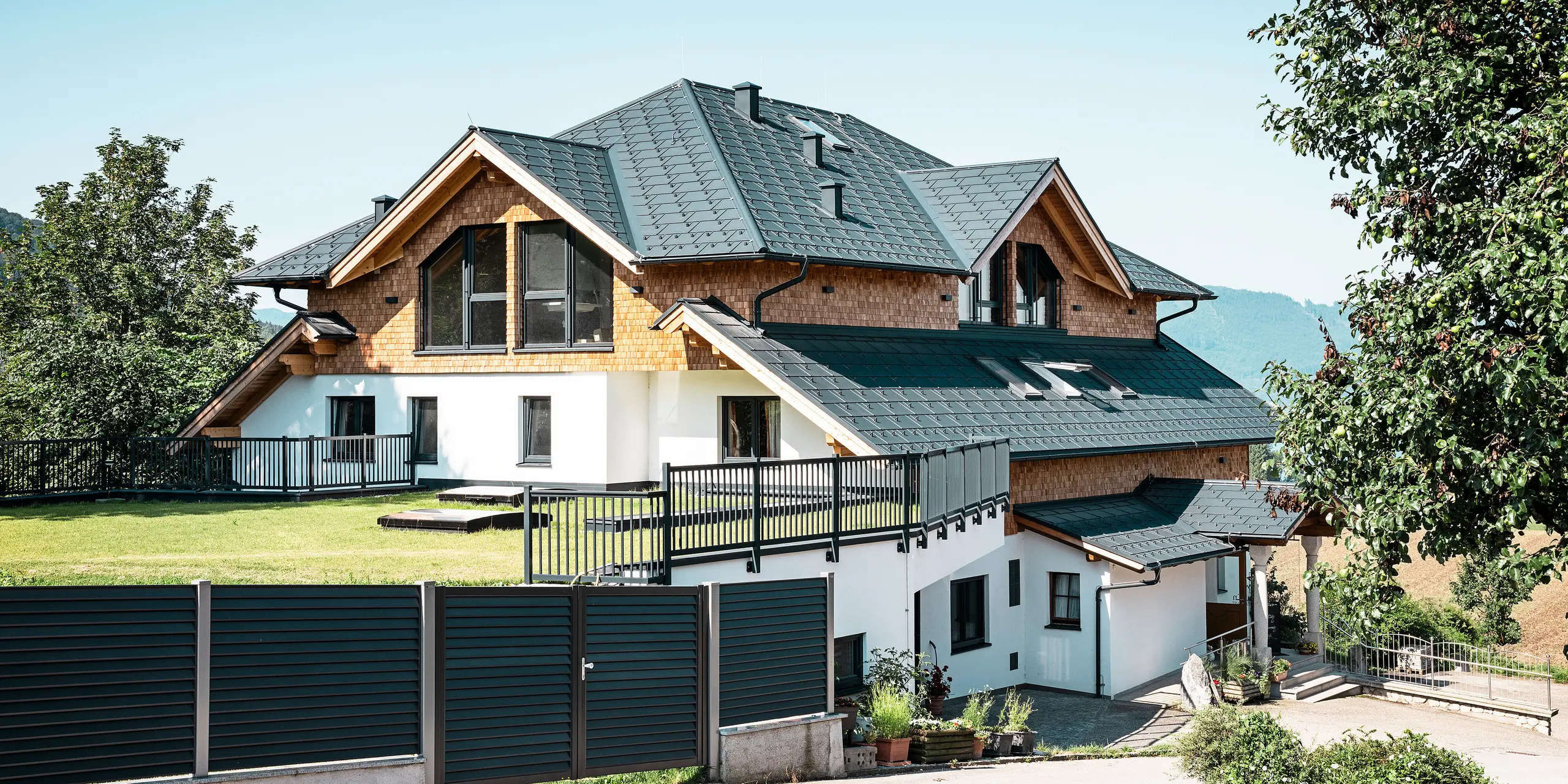 Bočný pohľad na kaviareň-reštauráciu Zenz'n Stub'n v Schörflingu pri jazere Attersee s modernou hliníkovou strechou zo strešných panelov PREFA R.16 vo vysokokvalitnej antracitovej farbe P.10. Odolná strecha PREFA spája estetiku a funkčnosť a poskytuje optimálnu ochranu. Prístup do záhrady a harmonické začlenenie budovy do prírodného prostredia podčiarkujú nadčasový dizajn.