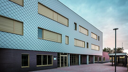Fotografia zobrazuje vstupnú stranu školy Collège Léontine Dolivet s modernou hliníkovou fasádou z fasádnych šablón PREFA v striebornom metalickom vyhotovení. Vzhľad budovy dotvárajú viaceré obdĺžnikové okná so žltými rámami a zatvorenými roletami. V popredí sa nachádza dláždené predsieňové priestranstvo, ktoré lemuje krytá časť. Fasáda pôsobí elegantne, je odolná voči poveternostným vplyvom a má dlhú životnosť.