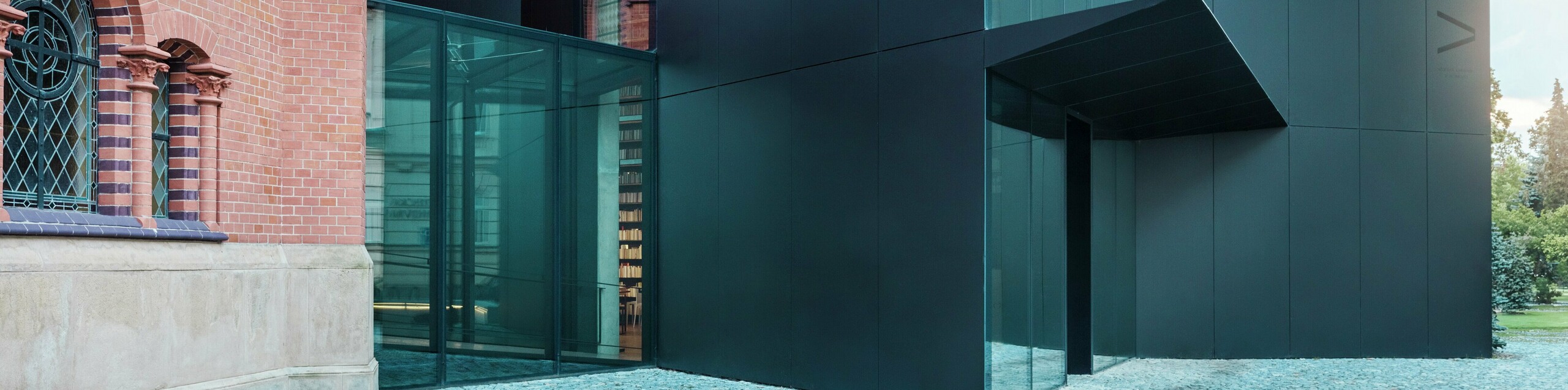 Obrázok ukazuje modernú prístavbu knižnice, ktorá elegantne nadväzuje na historický Červený kostol v Olomouci. Výrazná novostavba zaujme fasádou z čiernych kompozitných dosiek PREFABOND z hliníka, ktorá je navrhnutá v jasných geometrických tvaroch a presvedčí precíznou prácou s detailmi. Sklenené steny budovy ponúkajú transparentný pohľad do interiéru, ktorý harmonicky zapadá do historického prostredia. Súhra moderných materiálov a klasických architektonických prvkov vytvára zaujímavé vizuálne prepojenie medzi starým a novým. Inovatívne použitie dosiek PREFABOND zaisťuje nielen moderný vzhľad, ale aj dlhú životnosť a odolnosť, ktoré budovu pripravujú na budúcnosť.