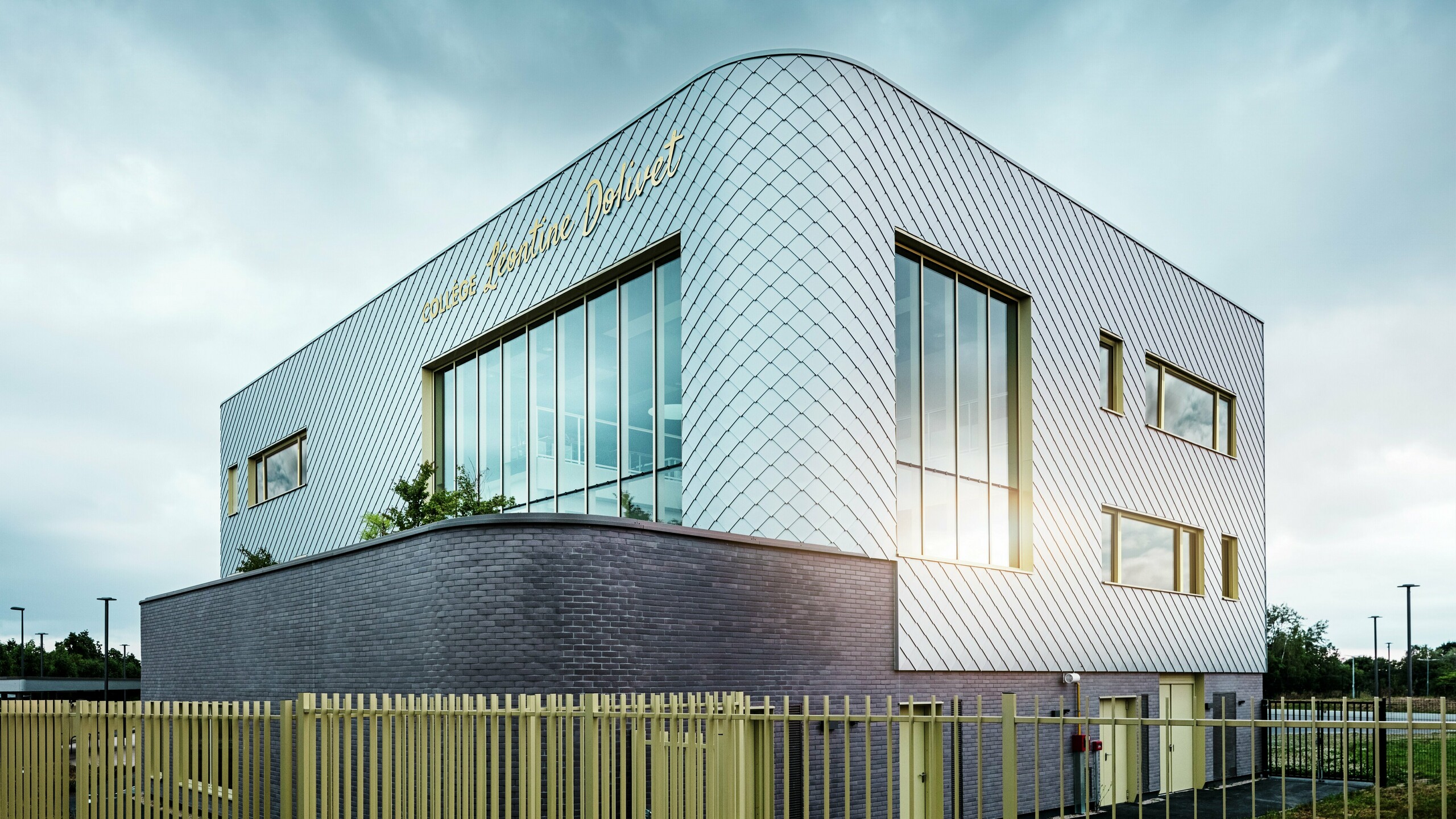 Fotografia zachytáva výrazný roh budovy Collège Léontine Dolivet s vlnitou hliníkovou fasádou zo strieborných fasádnych šablón. Veľké sklenené plochy odrážajú svetlo, zatiaľ čo žlté okenné rámy dodávajú budove akcenty. V popredí sa pozdĺž budovy tiahne svetlý plot, ktorý stojí na tmavom podstavci z tehál. Obloha je zamračená.