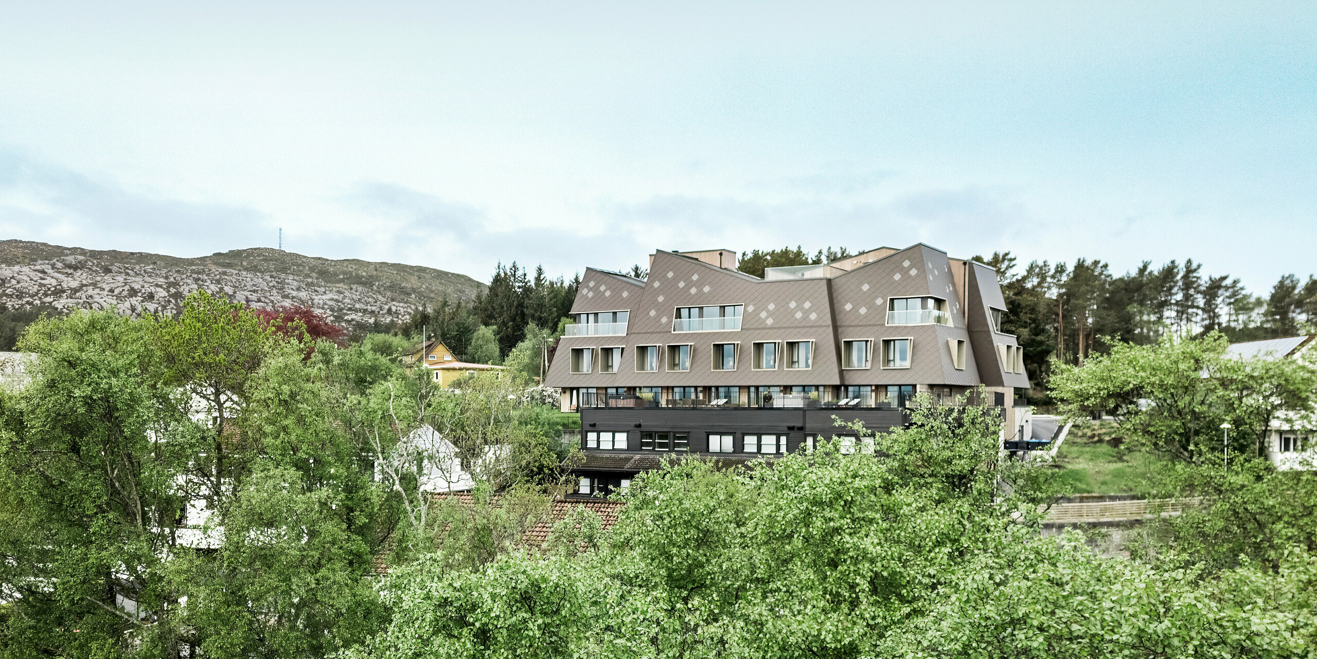 Pohľad z diaľky na butikový hotel Beckerwyc House v nórskom meste Bekkjarvik, ktorý je zasadený do zelenej kopcovitej krajiny. Futuristicky vyzerajúca budova je obložená fasádnou šablónou PREFA 44 × 44 v orieškovohnedej farbe, ktorá harmonicky zapadá do skalnatého okolia. Neobvyklá architektúra hotela sa odlišuje od okolitých tradičných domov, a napriek tomu vďaka zemitej farebnosti dobre splýva s prírodou.