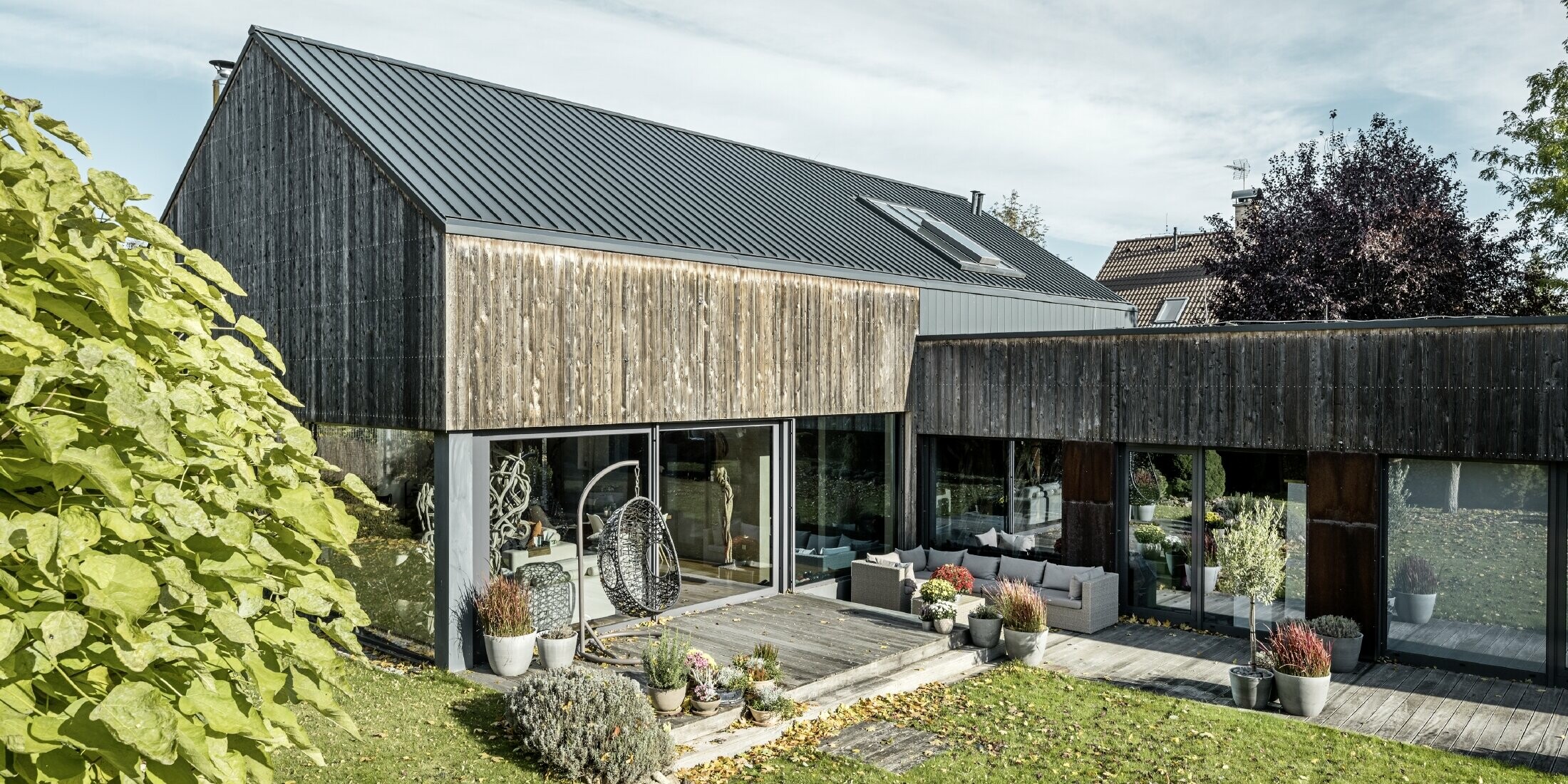 Rodinný dom so sedlovou strechou, zakrytou s dvojitou stojatou drážkou PREFALZ vo farebnom vyhotovení antracitová a zvetranou drevenou fasádou. S peknou drevenou terasou a veľkými okennými plochami na prízemí.