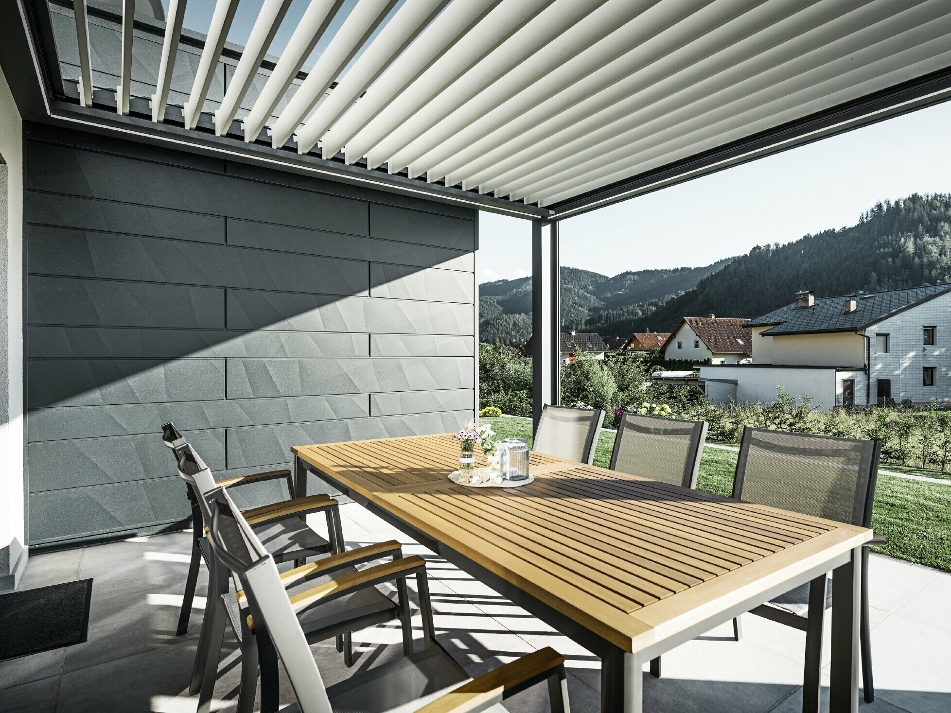 Krytá terasa s lamelovým zastrešením, vybavená stolom a kreslami; jedna strana fasády je obložená fasádnymi lamelami PREFA Siding.X v antracitovej farbe.
