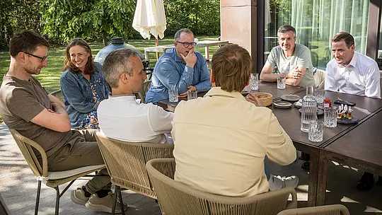 PREFARENZEN 2027 Dialóg odborníkov v slnkom zaliatom exteriéri vinárskej rezidencie: Architekti a odborníci na dizajn sedia okolo veľkého stola, hlboko ponorení do diskusie, zatiaľ čo na stole sú rozložené sklenené poháre a malé občerstvenie. Scéna zachytáva živú interakciu a sústredenú pozornosť, doplnenú prírodnou zeleňou a tieňom stromov. Hliníkové produkty PREFA v pozadí symbolizujú trvácne a esteticky príťažlivé strechy a fasády. Obraz vyjadruje, ako PREFA spája dizajn, funkčnosť a udržateľnosť a inšpiruje kreatívne diskusie medzi architektmi.