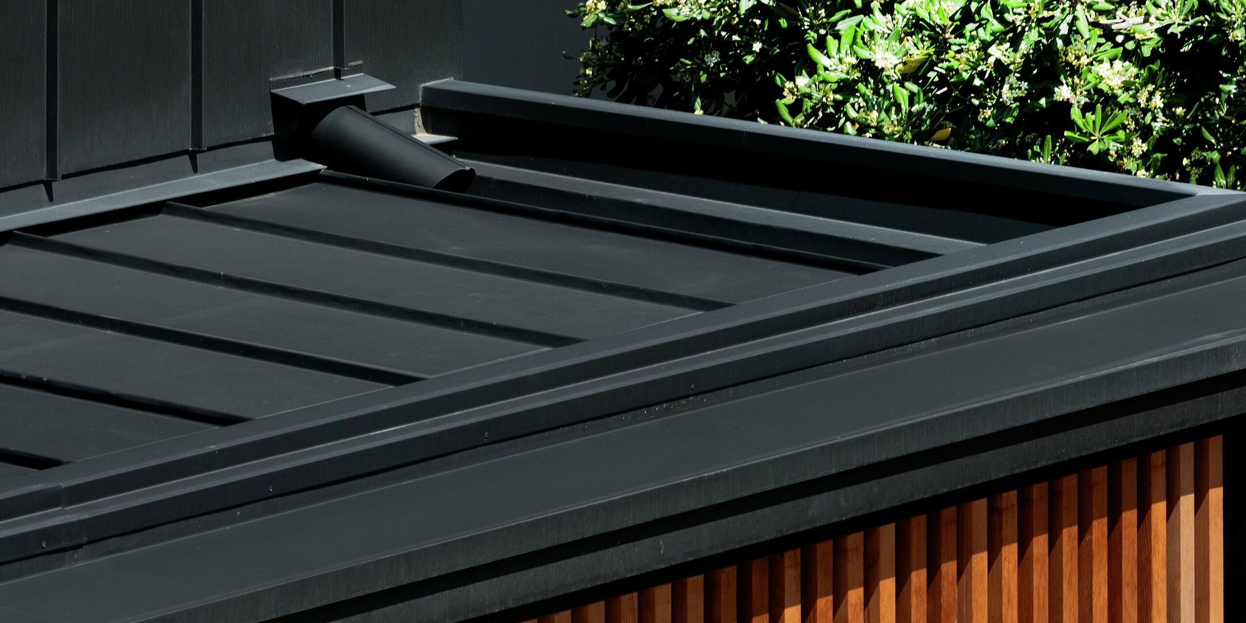 Detailansicht eines modernen Hauses in Cattolica: elegante Holzlamellenfassade kombiniert mit PREFALZ Dach und Fassade in Schwarzgrau. Die Aluminiumoberfläche von PREFA bietet Langlebigkeit, Korrosionsschutz und klare Linienführung – perfekt für zeitgemäße Architektur mit natürlichem Charme und hoher Widerstandsfähigkeit. Am Bild zu sehen ist die versteckte Dachentwässerung.