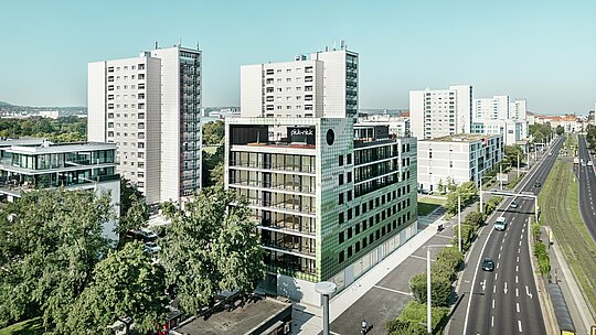 Letecký pohľad na obytnú a komerčnú budovu „Pick-Nick“ na Straßburger Platz v Drážďanoch s výraznou fasádou PREFA z hliníkových šablón v rôznych odtieňoch zelenej a béžovej farby. Budova je zasadená do moderného mestského prostredia s výškovými budovami a zelenými plochami a nachádza sa na hlavnej ceste. Výrazná architektúra a vysokokvalitná fasáda PREFA vytvárajú vizuálne akcenty v mestskej krajine.