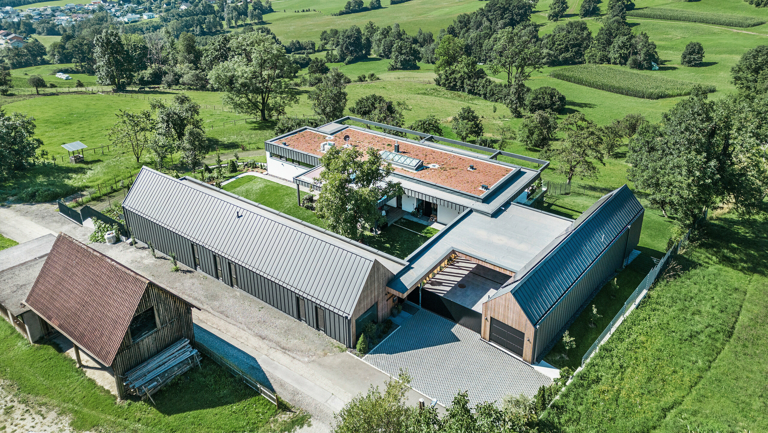 Letecký pohľad na priestranný rodinný dom kombinovaný s poľnohospodárskymi stajňami v rakúskom meste Pogier, zasadený do malebnej krajiny s výhľadom na zvlnené kopce a zelené polia. Budovy sú vybavené strešným a fasádnym systémom PREFALZ vo farbe tmavošedá P.10, ktorý vnáša do vidieckeho prostredia modernú estetiku. Usporiadanie budov vytvára príjemnú atmosféru dvora, zatiaľ čo fotovoltický systém na jednej zo striech odráža záväzok domu k udržateľnosti a energetickej účinnosti.