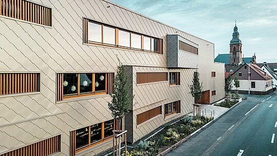 Bočný pohľad na detské jasle v Alzenau ukazuje modernú hybridnú budovu v napínavom kontraste s historickou štruktúrou mesta s neskorogotickým hradným kostolom v pozadí. Fasáda z bronzových PREFA šablón 44 × 44 vytvára elegantný, rytmický vzor a sebavedome vyniká z okolia – bez toho, aby pôsobila rušivo. Veľkorysé okenné otvory s vertikálnymi drevenými lamelami a štruktúrovaná zeleň pozdĺž budovy podčiarkujú udržateľný dizajnový koncept. Kombinácia inovatívnej hliníkovej fasády, ekologického spôsobu výstavby a citlivého prístupu k urbanistickému riešeniu robí z tohto projektu ukážkový príklad zodpovednej architektúry medzi tradíciou a budúcnosťou.