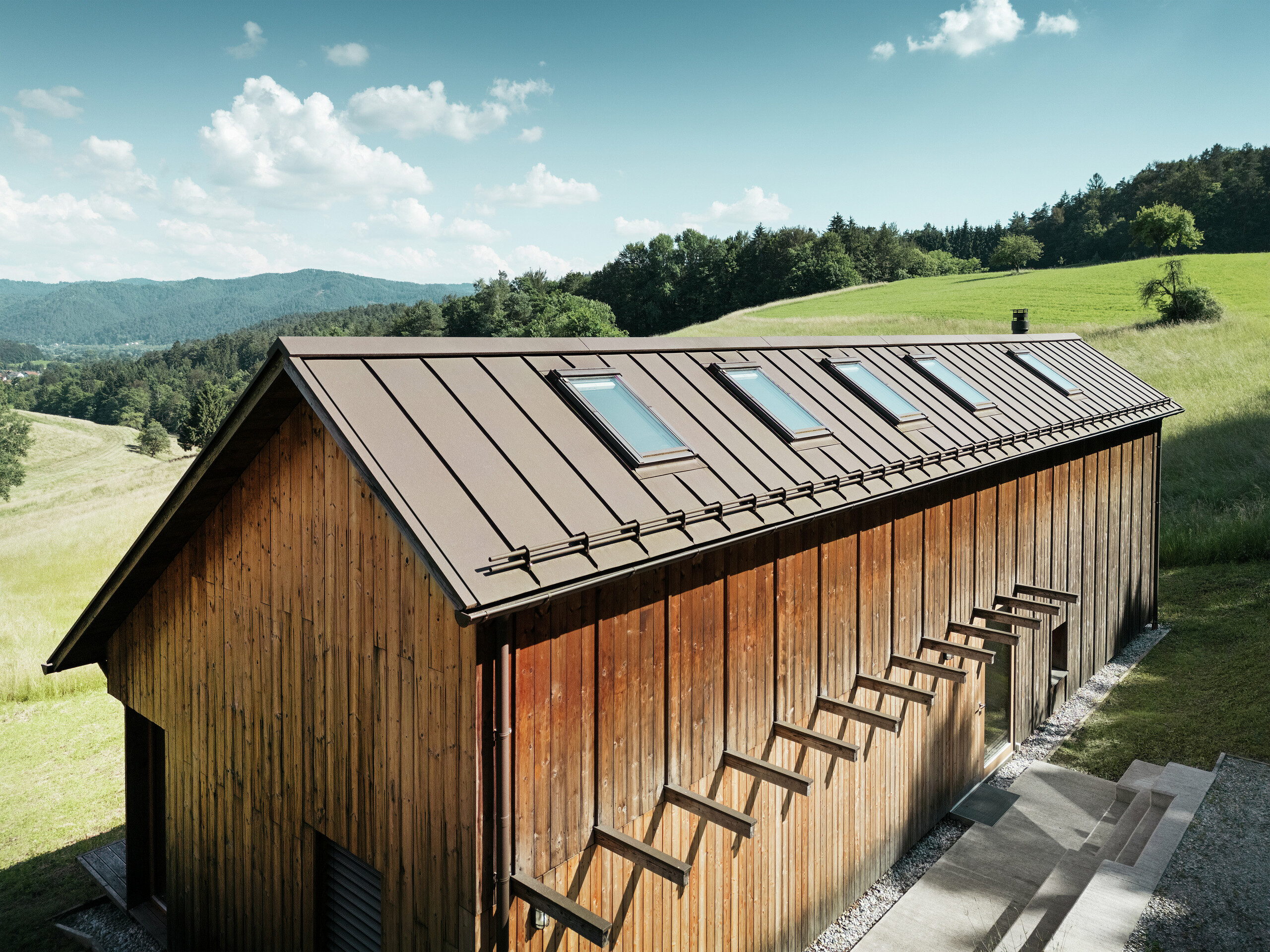 Architektonicky jasne štruktúrovaná drevostavba s kvalitnou hliníkovou strechou PREFALZ v hnedej farbe P.10. Strecha PREFALZ s nízkymi nárokmi na údržbu zaujme presnými líniami, vysokou odolnosťou proti UV žiareniu a korózii a dokonalým zabudovaním strešných okien. Kombinácia udržateľnej drevenej fasády a odolnej strešnej krytiny so stojatou drážkou harmonicky zapadá do idylickej kopcovitej krajiny. Asymetrická šikmá strecha má päť presne umiestnených strešných okien, ktoré zabezpečujú svetlo v interiéri.