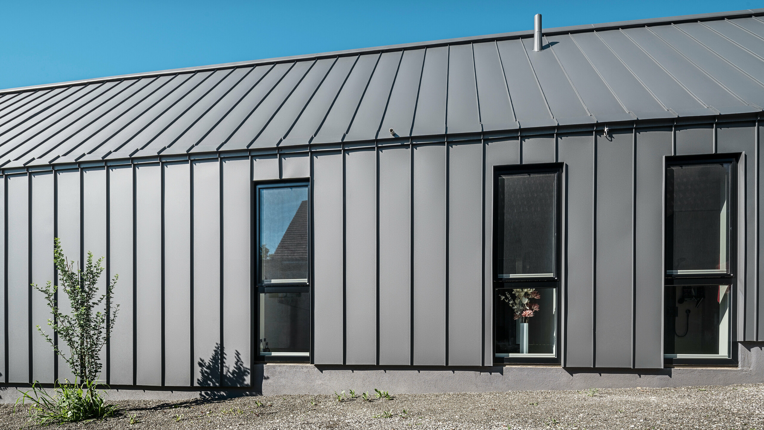 Bočný pohľad na rodinný dom v rakúskom Pogieri s odolným a elegantným zvitkovým plechom PREFALZ v P.10 tmavošedej na streche a fasáde. Hliníkový obklad so stojatou drážkou ponúka čisté vertikálne línie, ktoré elegantne rámujú okná a vytvárajú moderný vzhľad. Minimalistická architektúra stojí pod jasnou modrou oblohou.