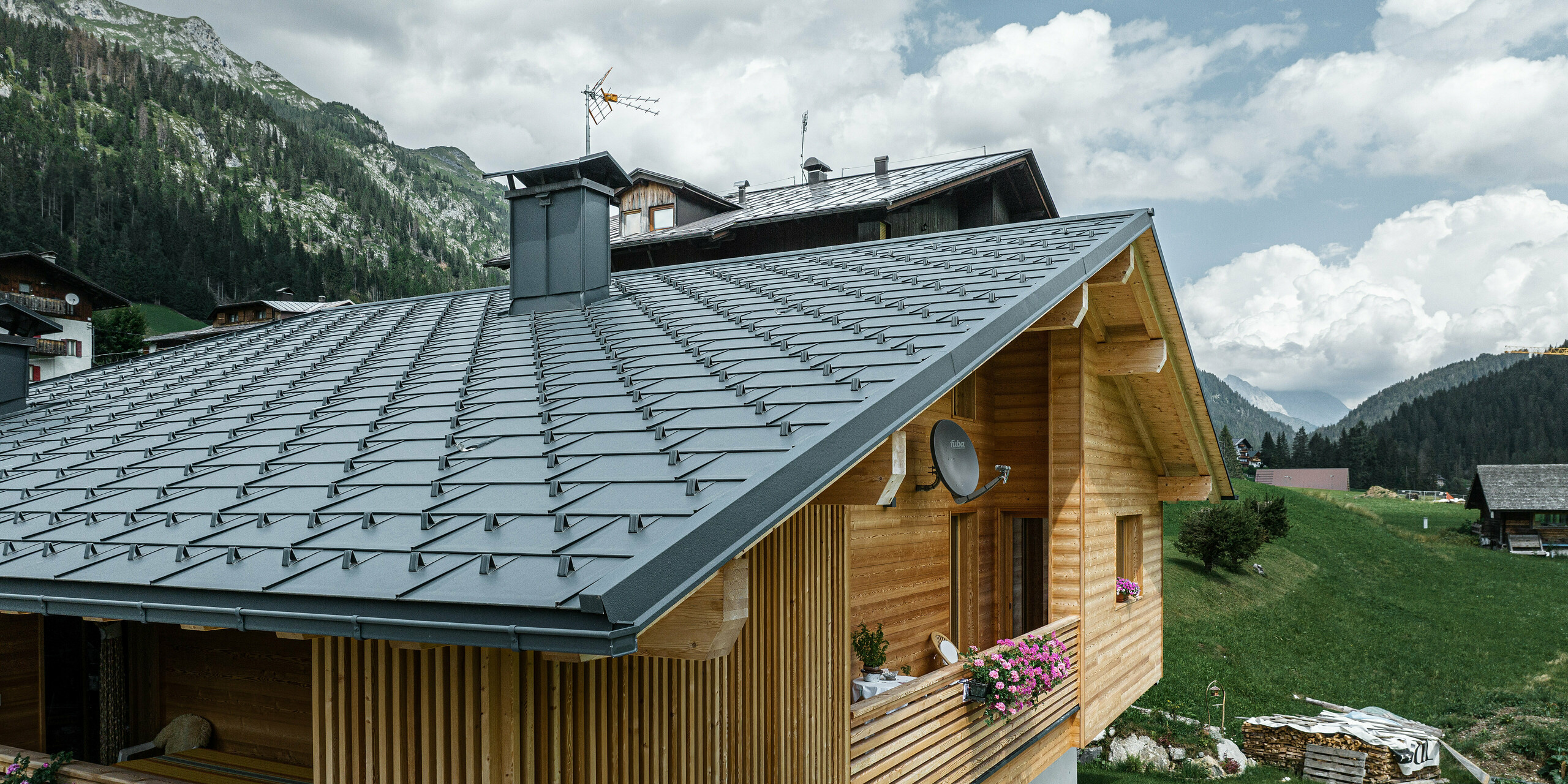 Perspektívny pohľad na Casa Hoffe v talianskej Sappade s odolnými strešnými panelmi PREFA R.16 v čiernej farbe P.10. Strecha bez problémov zapadá do tradičnej drevenej konštrukcie a ponúka estetický kontrast, ktorý dopĺňa alpská krajina. Kombinácia čierneho strešného systému PREFA a prírodnej drevenej fasády predstavuje symbiózu odolnosti, kvality a alpského šarmu.