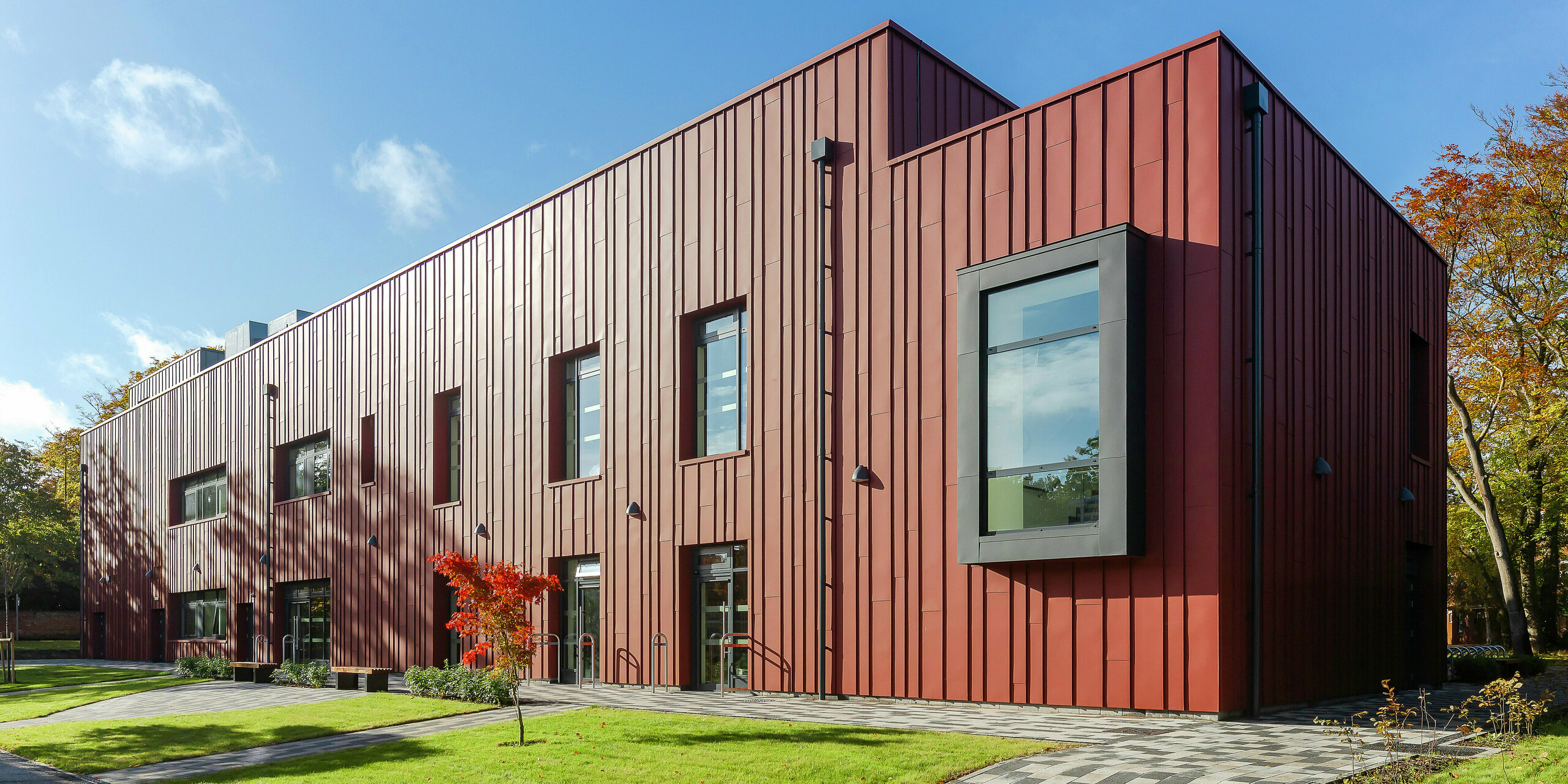 Moderná architektúra s robustnou hliníkovou fasádou: Soldiers' Centre v Pirbright sa pýši elegantnou stojatou falcovou krytinou PREFALZ v odtieni P.10 tmavočervená, ktorá harmonicky zapadá do zeleného prostredia. Výrazné, vysunuté okenné rámy v P.10 Anthrazit dodávajú architektúre osobitý charakter. Dláždená cesta a upravená zeleň podčiarkujú príjemný dizajn budovy. Udržateľné a estetické staviteľstvo s hliníkovými produktmi PREFA.