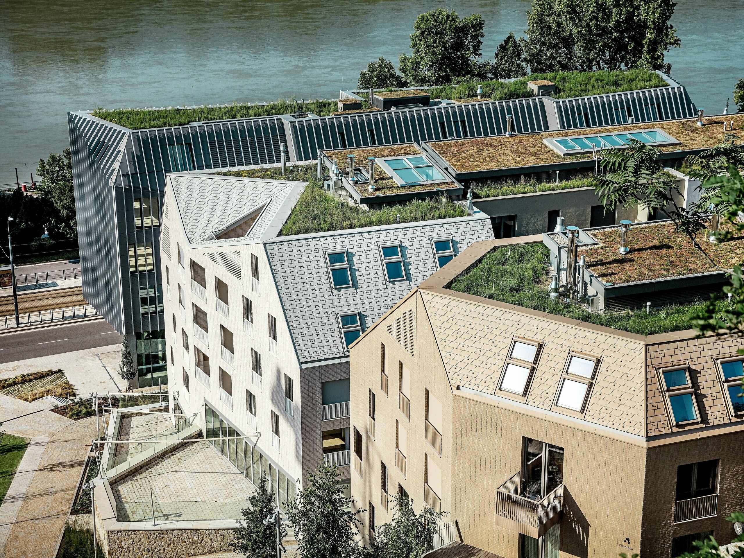 Moderná mestská štvrť v Bratislave s výraznou architektúrou a zelenými strechami priamo pri nábreží: Filigránske, strmo sklonené strechy a vikierové konštrukcie sú pokryté kvalitnými hliníkovými strešnými šindľami PREFA vo farbách prefa biela, pieskovo hnedá a bronzová, ktoré okamžite upútajú pozornosť. Veľkoformátové strešné okná, čisté línie a bohatá strešná zeleň prepájajú dizajn s udržateľnosťou. Odolné a bezúdržbové riešenia z hliníka PREFA presviedčajú pevnosťou, nízkou hmotnosťou a vysokou odolnosťou voči poveternostným vplyvom – ideálne pre náročné mestské stavebné projekty.