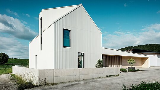 Moderný rodinný dom vo Freistadte z dynamického pohľadu, ktorý zdôrazňuje čisté línie a nápadnú architektúru. Čisto biela hliníková obkladová doska PREFALZ sa plynule tiahne po celej fasáde a streche a dodáva domu modernú a nadčasovú estetiku. Charakteristickými prvkami návrhu sú strmá sedlová strecha, veľká okenná plocha a výrazná vikierová strecha, zatiaľ čo vertikálne drážky fasády dodávajú budove zaujímavú textúru a hĺbku. Kontrast s prírodným prostredím a elegantné drevené prvky vo vstupnej časti a na garážových dverách dopĺňajú dizajn. PREFALZ zaisťuje mimoriadne odolný, ľahko udržiavateľný a poveternostne odolný povrch. Vďaka tomu je dom nielen esteticky príťažlivý, ale aj funkčný a udržateľný.