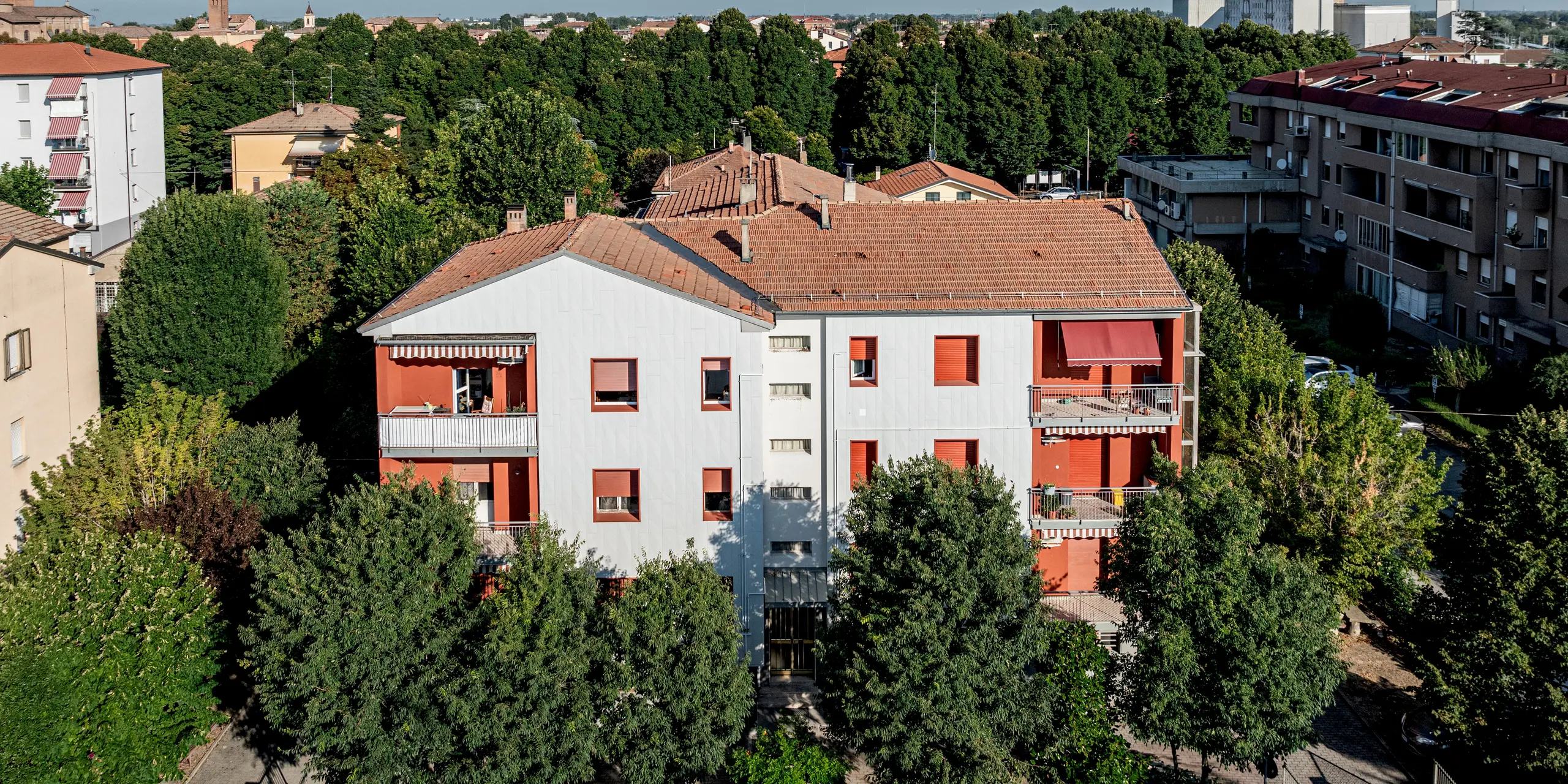 Weitblick über die Wohnanlage Alba B inmitten einer grünen, gewachsenen Stadtstruktur: Das sanierte Gebäude hebt sich mit seiner hellen PREFA Fassade aus Siding.X Aluminiumpaneelen deutlich von den umliegenden Ziegeldächern ab. Die vertikal verlegten Paneele sorgen für eine feine, strukturierte Oberfläche, während rote Fensterrahmen und Balkone prägnante Akzente setzen. Umgeben von dichtem Baumbestand wirkt das Ensemble lebendig und modern. Die PREFA Aluminiumfassade überzeugt durch ihre Langlebigkeit, Witterungsbeständigkeit und den geringen Wartungsaufwand.