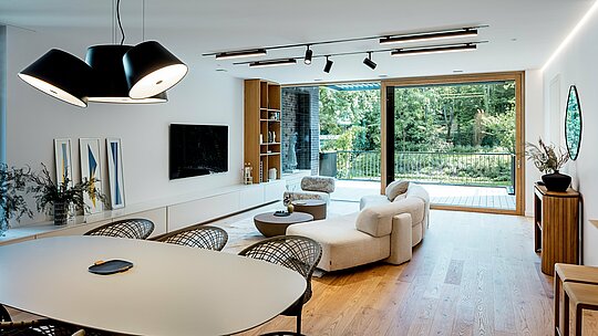 Záber zachytáva svetlom naplnený obývací a jedálenský priestor modernej viacgeneračnej vily v Budapešti. Veľké presklené plochy otvárajú interiér smerom k zelenej terase, zatiaľ čo svetlé drevené podlahy, elegantný dizajnový nábytok a harmonicky zladené osvetlenie vytvárajú príjemnú atmosféru. Otvorená dispozícia plynulo prepája kuchyňu, jedálenský kút a obývaciu izbu. Kvalitný interiérový dizajn dopĺňa čistý architektonický výraz budovy, ktorej vonkajšia štruktúra je definovaná trvácnymi hliníkovými produktmi PREFALZ v odtieni P.10 antracit. Priestor vyžaruje veľkorysosť, modernosť a vysoký komfort bývania.