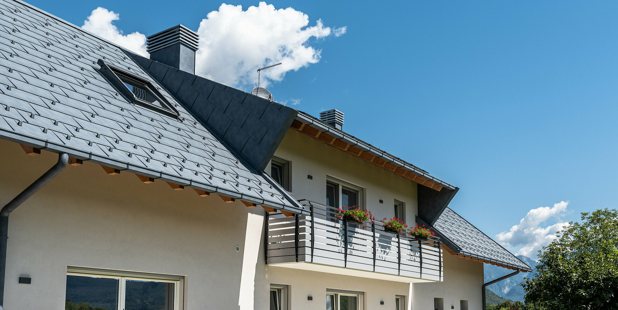 Pohľad zdola na elegantnú strechu domu v Belluno v Taliansku. Strešná plocha má strešné okno a bola pokrytá strešnými panelmi PREFA R.16 vo farbe P.10 antracitová. Strešné odvodnenie pozostávajúce z hranatého žľabu a odtokovej rúry je farebne zladené s robustnou hliníkovou strechou a vytvára jednotný a moderný vzhľad s optimálnou ochranou.