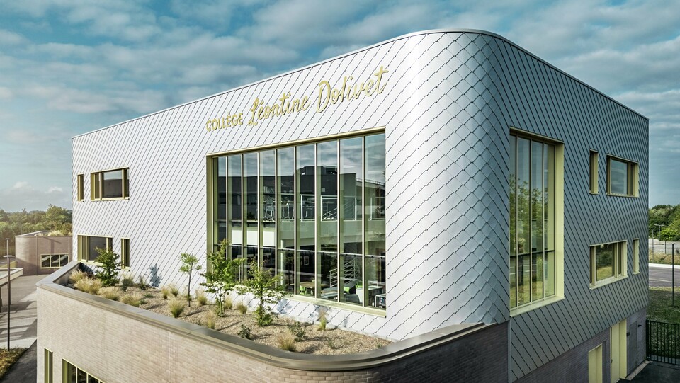 Fotografia zachytáva výrazný roh budovy Collège Léontine Dolivet v Cesson-Sévigné. Fasáda je tvorená striebornými hliníkovými fasádnymi šablónami PREFA, ktoré sú usporiadané do diagonálneho vzoru a odrážajú svetlo. Pozornosť upúta zakrivený tvar horného poschodia, ktorý budove dodáva jemný, moderný vzhľad. Veľké sklenené fasády otvárajú pohľad do vnútra, zatiaľ čo žlté okenné rámy dodávajú živé akcenty. V popredí sa nachádza ozelenená plocha so štrkom a kríkmi, ktorá harmonicky dopĺňa architektúru. Obloha je mierne zamračená, čo zdôrazňuje kovový povrch.