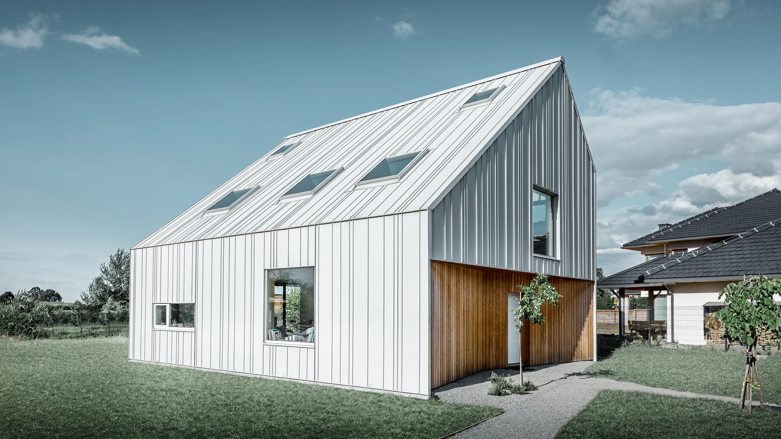 Moderne razený rodinný dom so sedlovou strechou, kompletný vonkajší plášť je obložený s PREFALZ vo farebnom vyhotovení prefa biela Pritom sa rozdielne široké pásy ťahajú od strechy ďalej cez fasádu. V streche sú zabudované viaceré strešné okná.