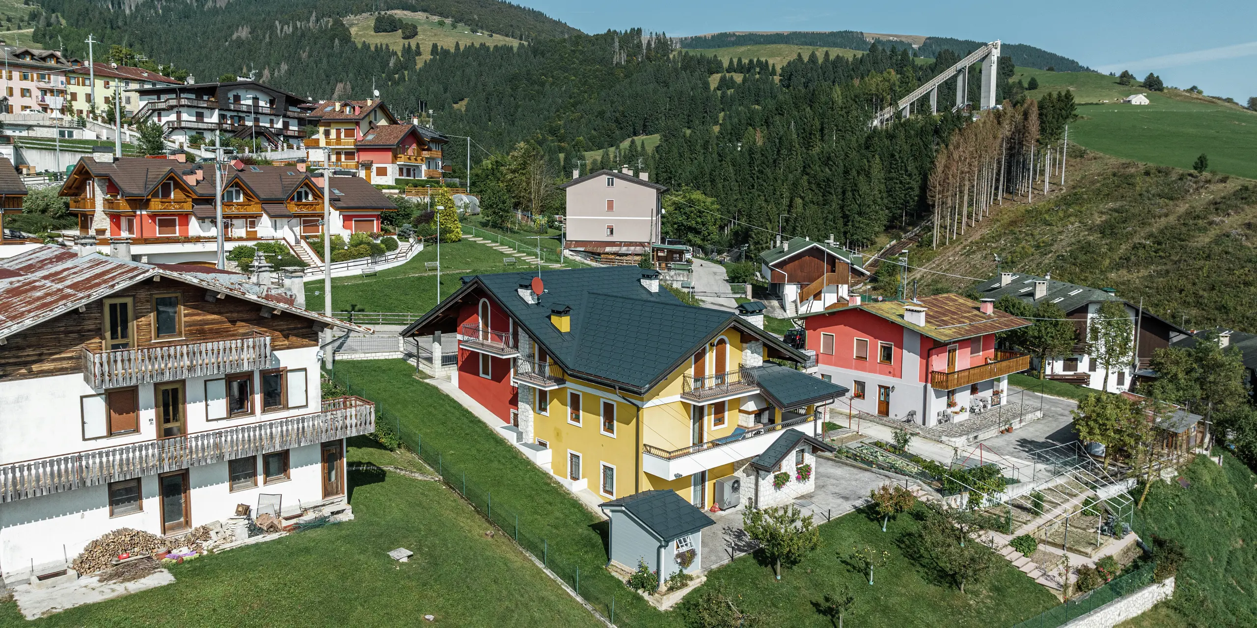 Idylický rodinný dom v Gallio s modernou plechovou strechou PREFA zo strešných panelov R.16 v tmavošedej farbe P.10. Odolná strešná krytina z hliníka spoľahlivo chráni dom pred snehom, dažďom a vetrom – ideálna pre alpské regióny. Žlto-červená fasáda s akcentmi z prírodného kameňa harmonicky zapadá do zeleného prostredia. Husté lesy a skokanský mostík v pozadí podčiarkujú typický alpský charakter regiónu. Trvanlivá, odolná a estetická – perfektná strecha pre celé generácie.