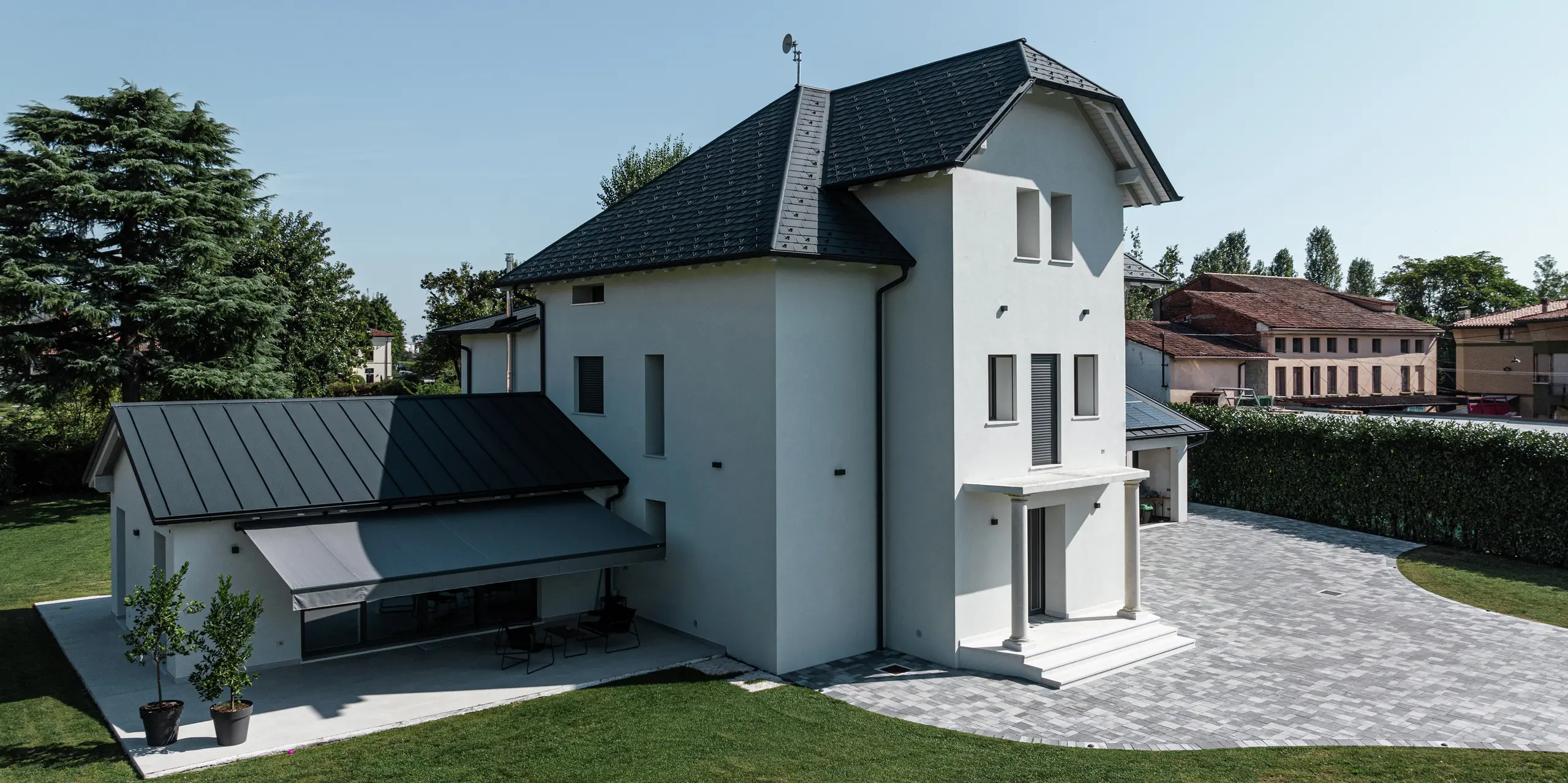 Bočný pohľad na moderný rodinný dom v Marano Vicentino v Taliansku s robustnými strešnými šindľami PREFA v antracitovej farbe P.10. Priľahlá vedľajšia budova bola vybavená odolnou dvojitou stojatou falcovou krytinou. Svetlosivá markíza poskytuje tieň v čase silného poludňajšieho slnka. Čistá architektúra a vysoko kvalitné hliníkové produkty zaisťujú moderný a odolný strešný povrch.