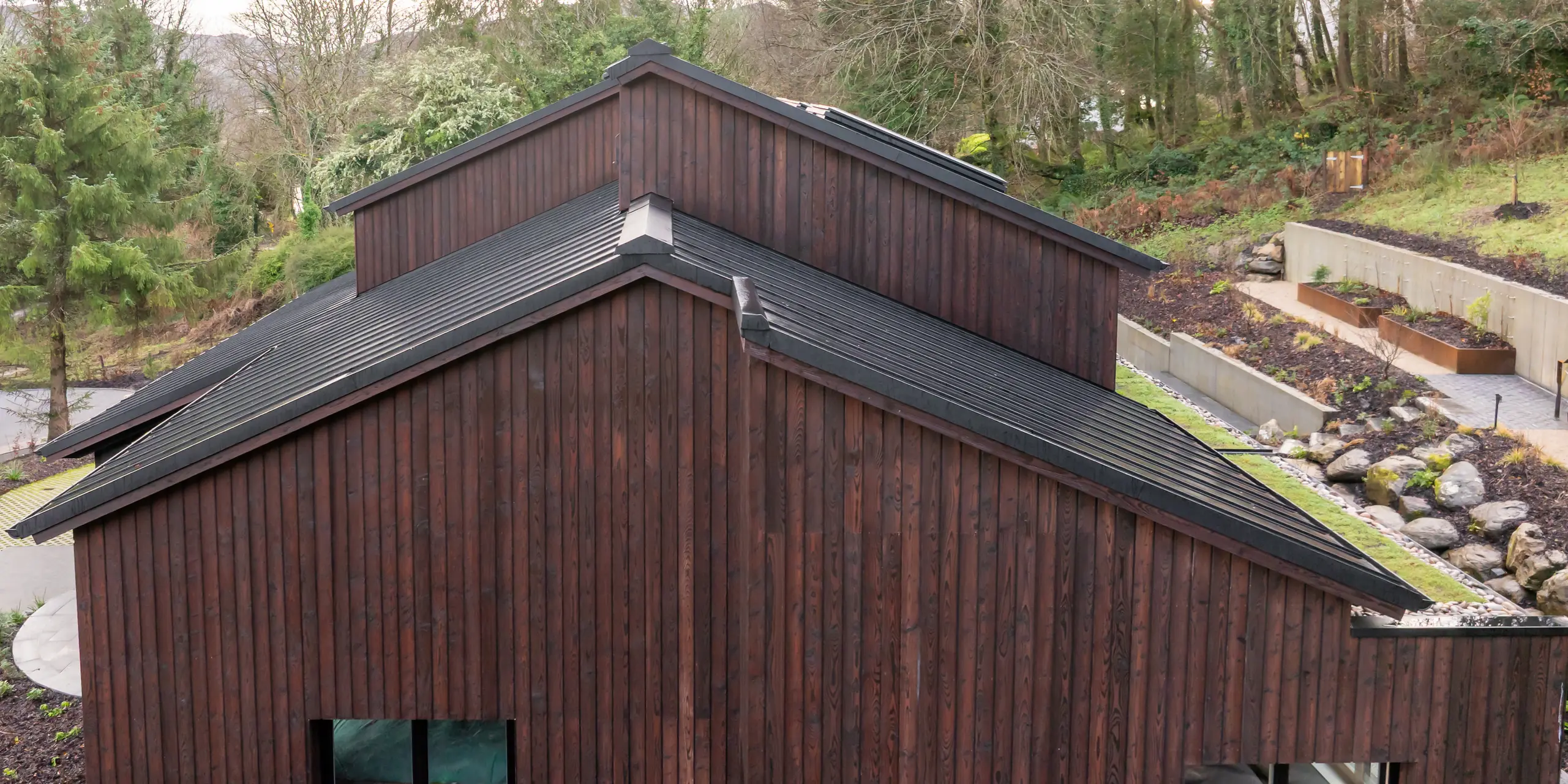 Seitenansicht des House of Holywell in Sligo: Die markanten Giebelformen führen den Blick entlang der klar geschnittenen Dachlinien, die vom PREFALZ Aluminiumdach in P.10 Schwarz präzise nachgezeichnet werden. Die feinen Stehfalze strukturieren die geneigten Flächen und unterstreichen die moderne Architektursprache. In Kombination mit der warmen Holzfassade entsteht ein starker Kontrast. Das PREFA Dach überzeugt durch Langlebigkeit, Korrosionsbeständigkeit und zuverlässigen Schutz bei anspruchsvollen Wetterbedingungen.