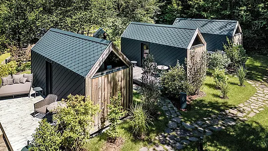 Letecký pohľad na niekoľko minimalistických minidomov v Secret Koi Garden pri jazere Natterer See, zasadených do hustej zelene a prírodných záhradných chodníkov. Strešné a fasádne plochy sú kompletne obložené hliníkovými strešnými a fasádnymi šablónami PREFA v odtieni P.10 antracit, ktoré svojou rovnomernou štruktúrou pripomínajúcou rybiu kožu dotvárajú celkový vzhľad. Ľahký, odolný a poveternostne odolný hliník PREFA umožňuje precízne detaily a podčiarkuje čistú architektúru glampingu v súlade s prírodou.