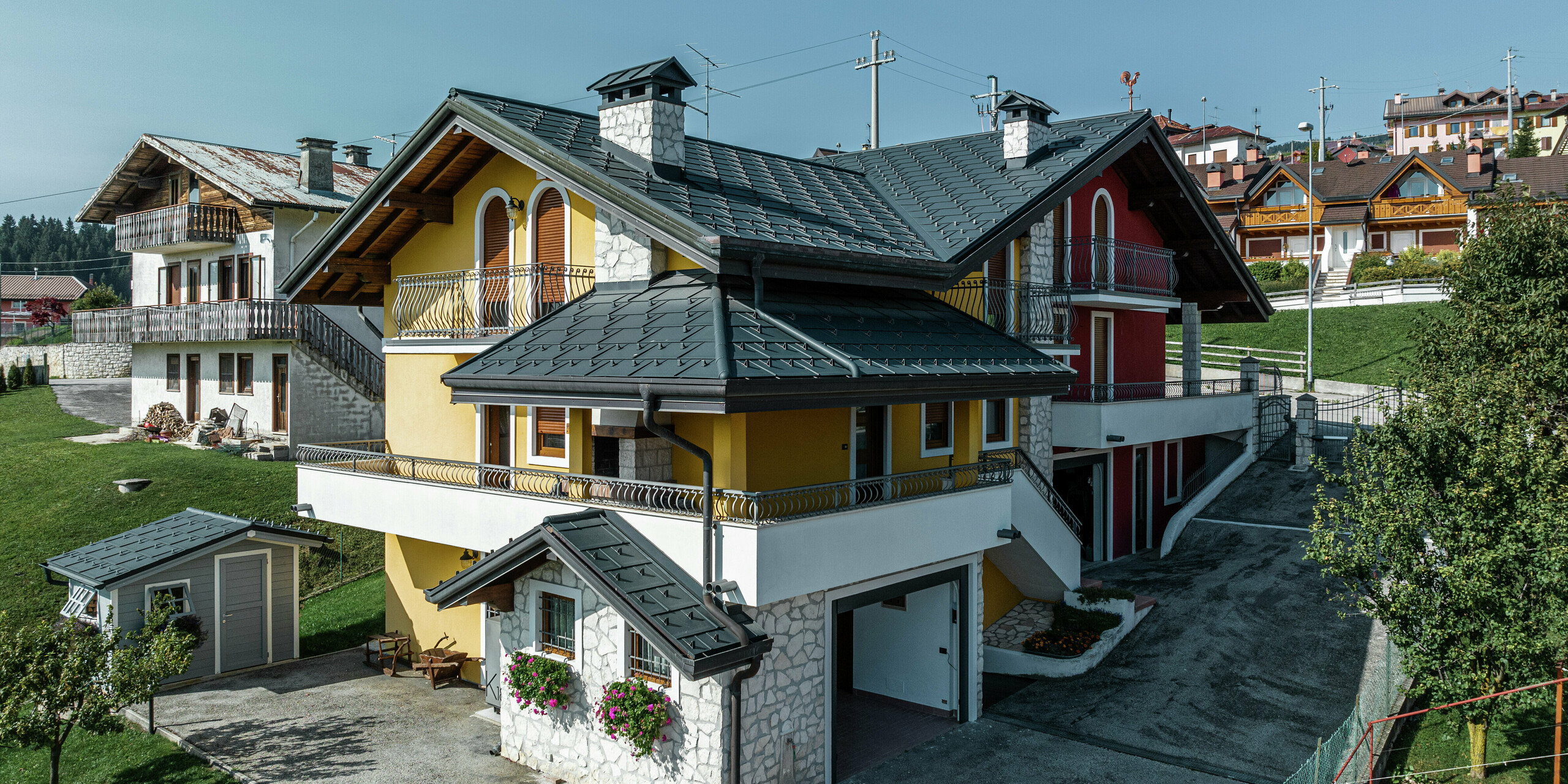 Rodinný dom v Gallio v alpskom architektonickom štýle s modernou plechovou strechou PREFA v tmavošedej farbe P.10. Strešné panely PREFA R.16 zaručujú trvácne, odolné voči poveternostným vplyvom a vizuálne atraktívne riešenie strechy. Harmonická kombinácia žlto-červenej fasády, prvkov z prírodného kameňa a drevených akcentov dodáva domu príjemný charakter. PREFA odvodnenie strechy v tmavošedej farbe P.10 zaisťuje spoľahlivé odvádzanie vody. Ukážkový príklad estetickej a funkčnej architektúry v alpskom prostredí.