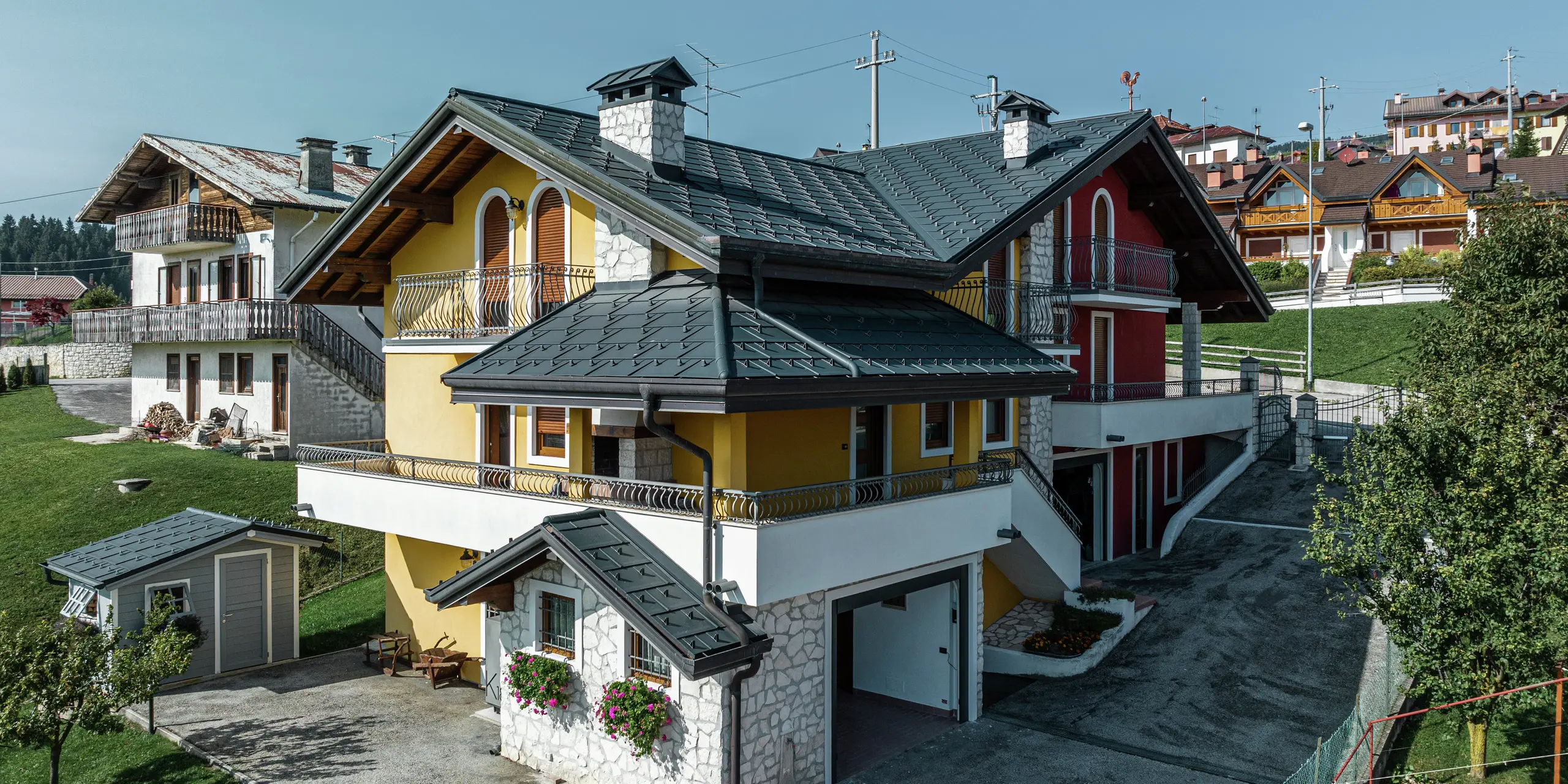 Rodinný dom v Gallio v alpskom architektonickom štýle s modernou plechovou strechou PREFA v tmavošedej farbe P.10. Strešné panely PREFA R.16 zaručujú trvácne, odolné voči poveternostným vplyvom a vizuálne atraktívne riešenie strechy. Harmonická kombinácia žlto-červenej fasády, prvkov z prírodného kameňa a drevených akcentov dodáva domu príjemný charakter. PREFA odvodnenie strechy v tmavošedej farbe P.10 zaisťuje spoľahlivé odvádzanie vody. Ukážkový príklad estetickej a funkčnej architektúry v alpskom prostredí.