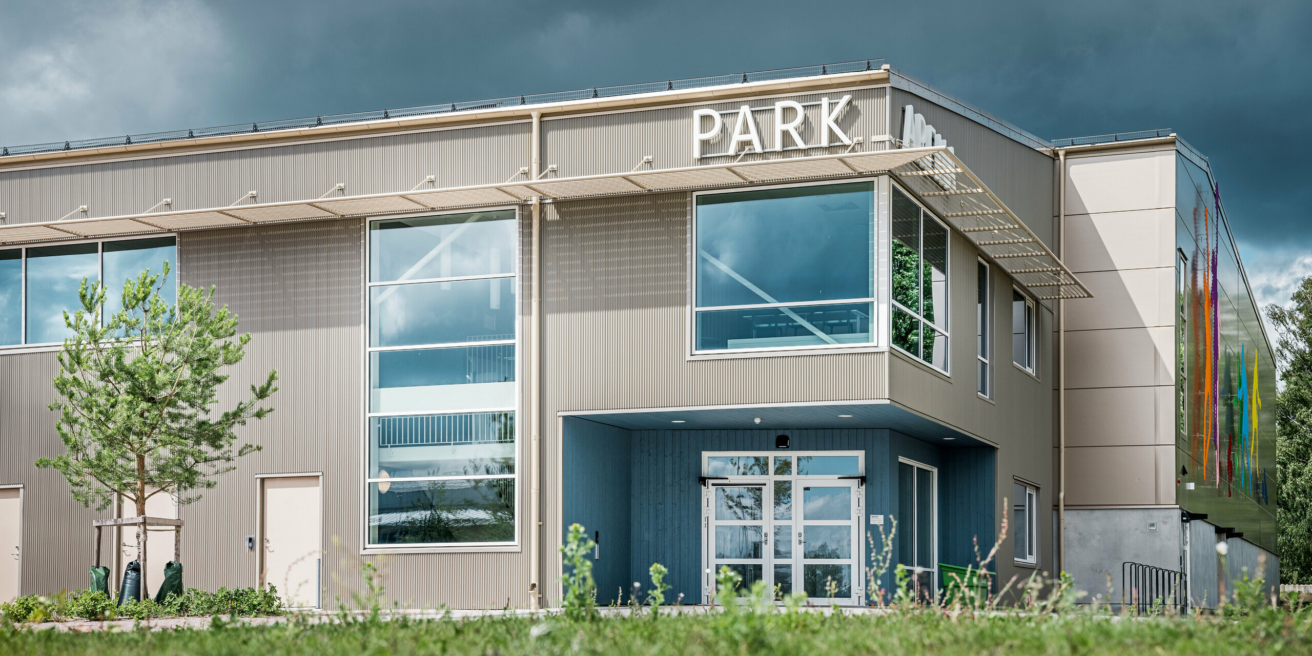 Vstupný areál Park Areny vo švédskom meste Värnamo z pohľadu žaby. Fasáda je obložená výrazným prelamovaným profilom PREFA v bronzovej farbe, ktorý vytvára elegantné efekty svetla a tieňa. Moderné sklenené priečelia a umelecky stvárnené figúry na plášti budovy zdôrazňujú inovatívny dizajn športovej haly