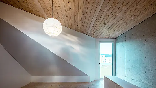 Obrázok zobrazuje minimalistický interiér rodinného domu vo Freistadte. Drevený strop z bieleho smreka dodáva miestnosti teplú a príjemnú atmosféru, ktorú podčiarkuje jednoduchý dizajn a prirodzené svetlo prúdiace oknom na pravej strane. Okno ponúka výhľad na zelené okolie a umocňuje pocit otvorenosti. Priestor je charakteristický hladkými, neutrálnymi stenami. Kombinácia betónovej steny a dreveného stropu vytvára harmonický kontrast, ktorý vyžaruje eleganciu aj prirodzenosť. Premyslený dizajn interiéru odráža filozofiu celého domu: funkčnosť sa snúbi s estetickou čistotou a nadčasovými materiálmi.