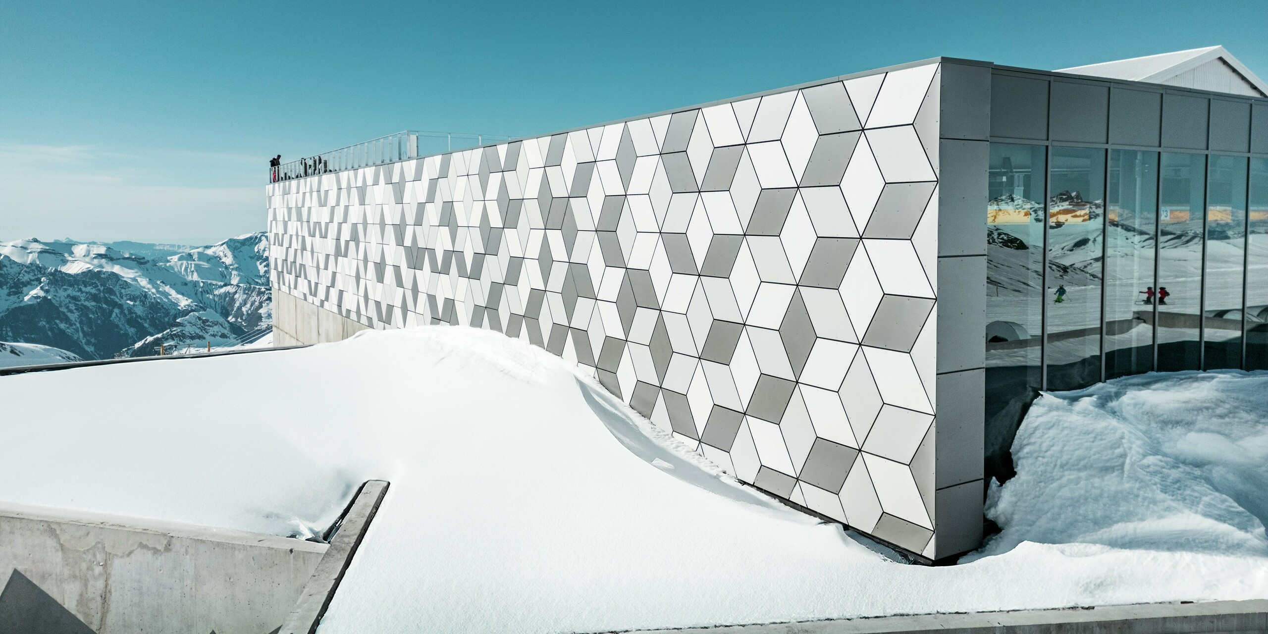 Horná stanica lanovky v Les Deux Alpes sa prezentuje úchvatnou fasádou z hliníkových kompozitných panelov PREFABOND usporiadaných do pútavého diamantového vzoru. Panely v bielej a sivej farbe dokonale zapadajú do zasneženej horskej krajiny a odrážajú jasné alpské svetlo. Tento inovatívny dizajn fasády v sebe spája estetickú prepracovanosť a vysokú odolnosť voči extrémnym poveternostným podmienkam.