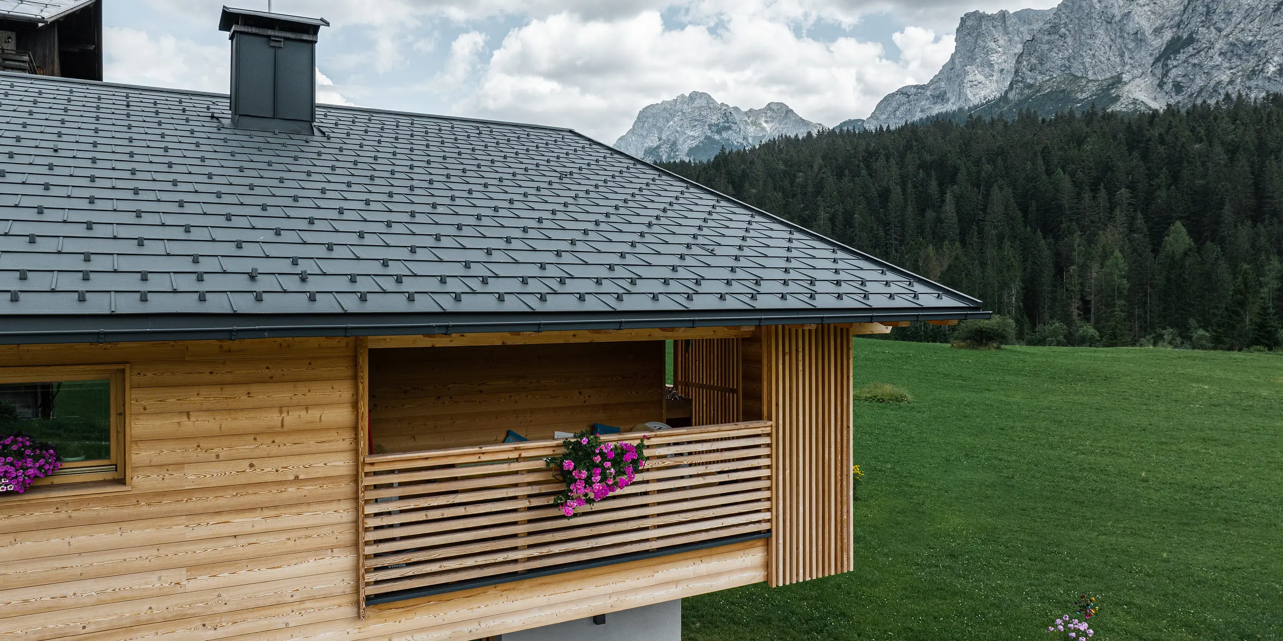 Malebný bočný pohľad na lodžiu Casa Hoffe v talianskej Sappade ukazuje harmonickú integráciu strešných panelov PREFA R.16 v čiernej farbe P.10 s prírodnou drevenou fasádou. Tradičná konštrukcia lodžie v kombinácii s moderným, odolným riešením strechy hladko zapadá do úchvatnej horskej kulisy Dolomitov a podčiarkuje prirodzený koncept bývania regiónu.