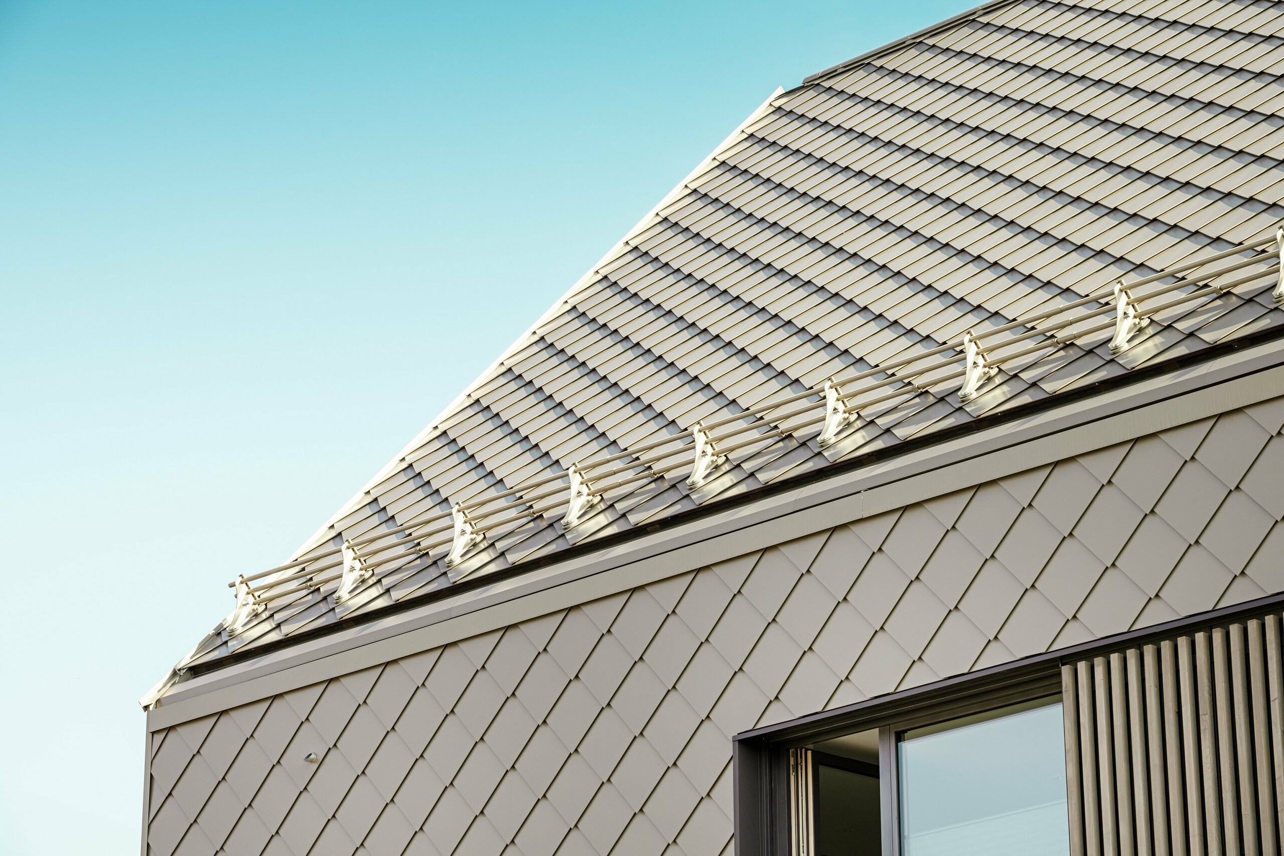 Detailný pohľad na bytový dom v Leitershofene, Nemecko, so strechou PREFA a fasádnymi šablónami 29 × 29 v bronzovej farbe. Povrch strechy je vybavený protisnehovou ochranou PREFA, ktorá zabezpečuje bezpečnosť pred padajúcimi strešnými lavínami. Hliníkové šablóny poskytujú trvanlivý povrch odolný voči poveternostným vplyvom a zdôrazňujú moderný dizajn.